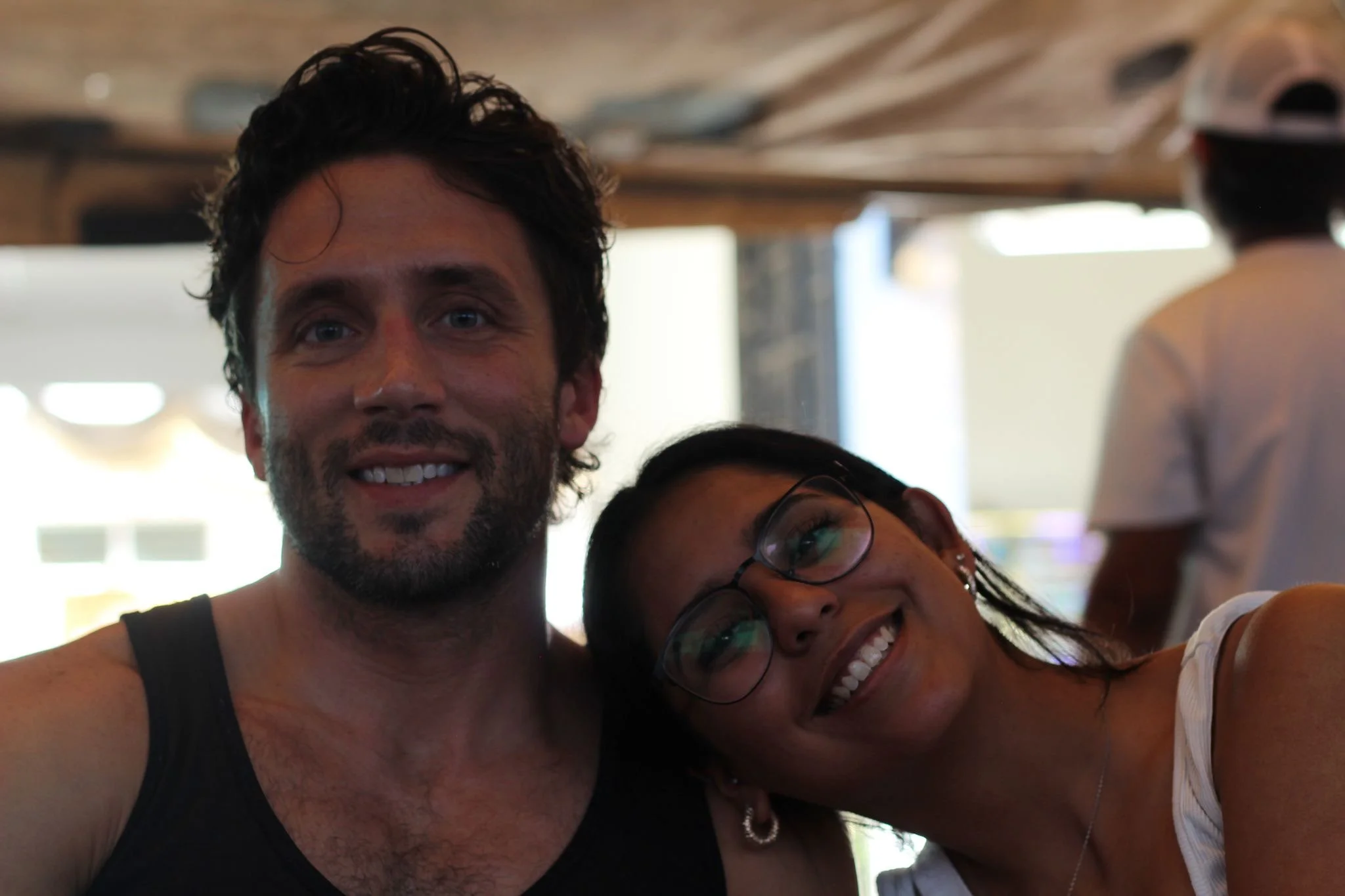 Two friends smiling and hugging each other indoors. The person on the left is a man with dark, curly hair, beard, and wearing a black tank top. The person on the right is a woman with glasses, long dark hair, wearing earrings, and a white top.