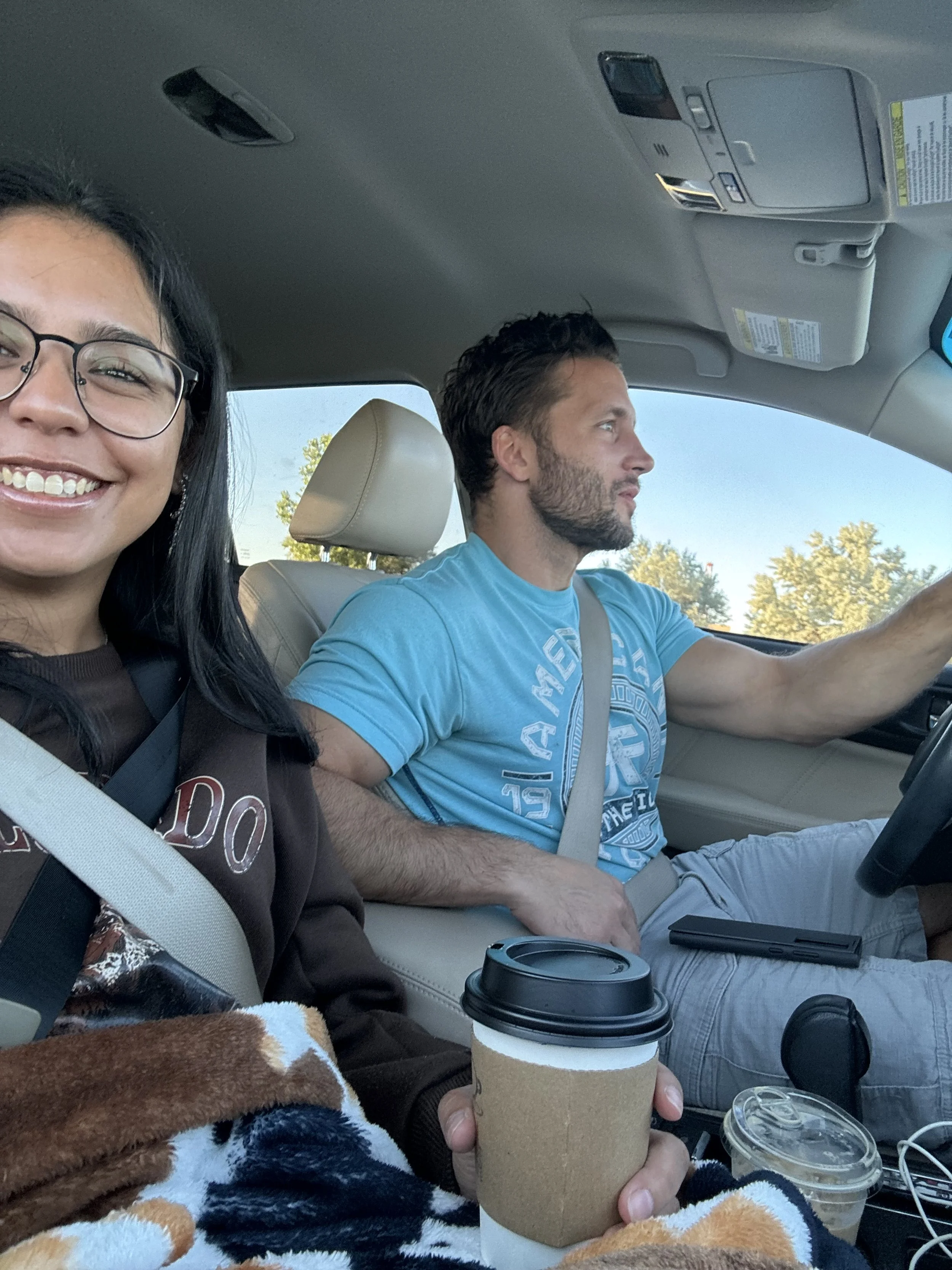 A woman with glasses smiling in the front passenger seat and a man driving in the front seat of a car. The woman is holding a to-go coffee cup and there is another coffee cup and some other items on her lap. Trees are visible outside the car window.