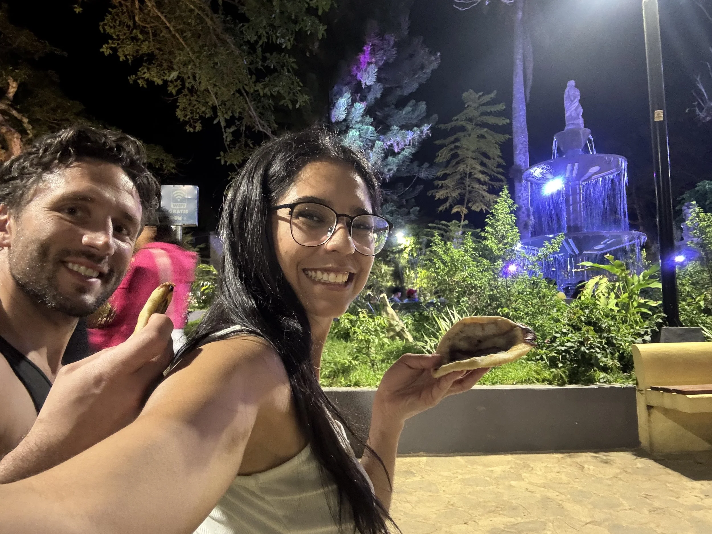 Two smiling people, a man and a woman, are holding large pretzels at an outdoor nighttime event. In the background, there is a water fountain, trees, and bright lighting.