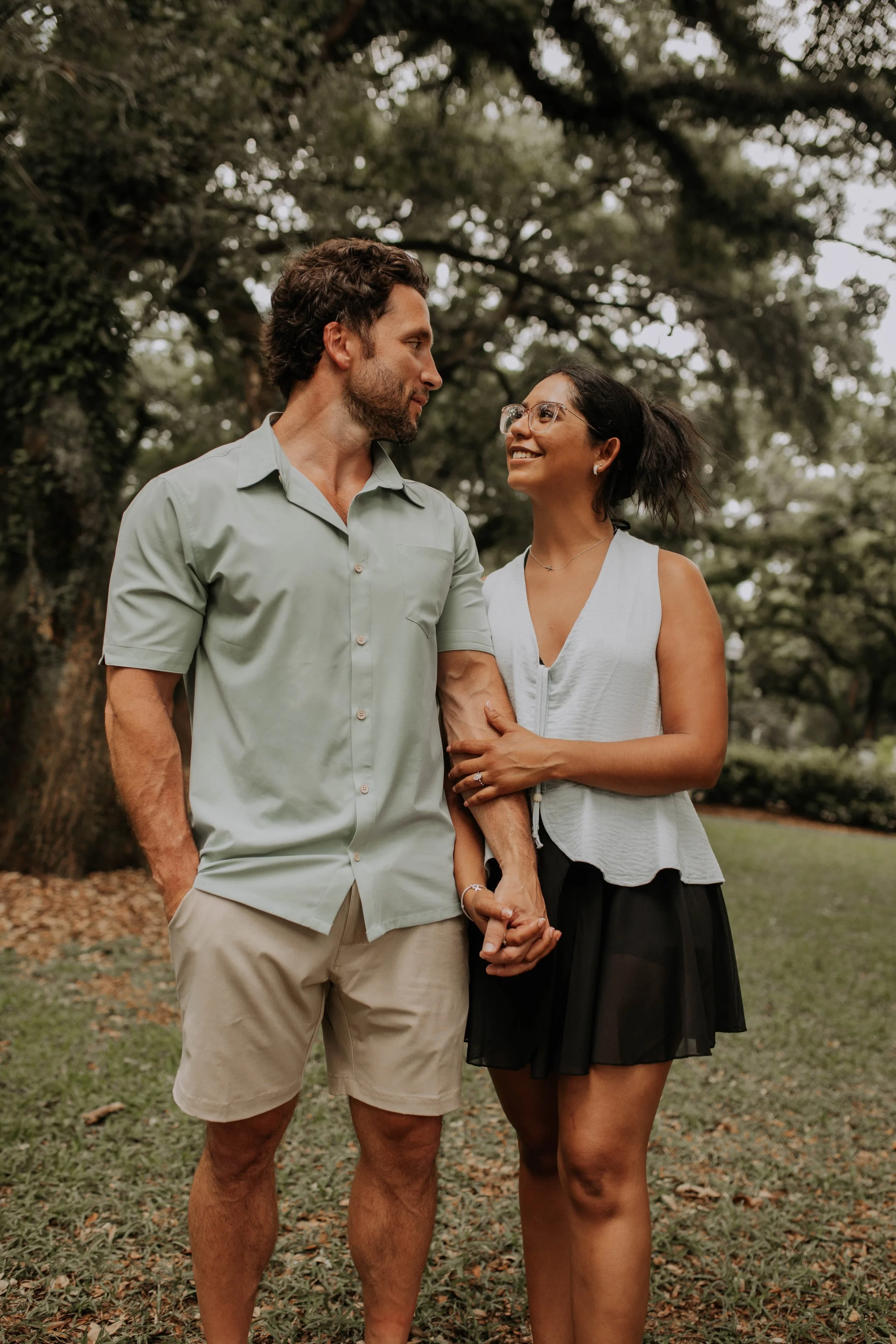A couple holding hands and smiling at each other outdoors in a park with trees in the background.