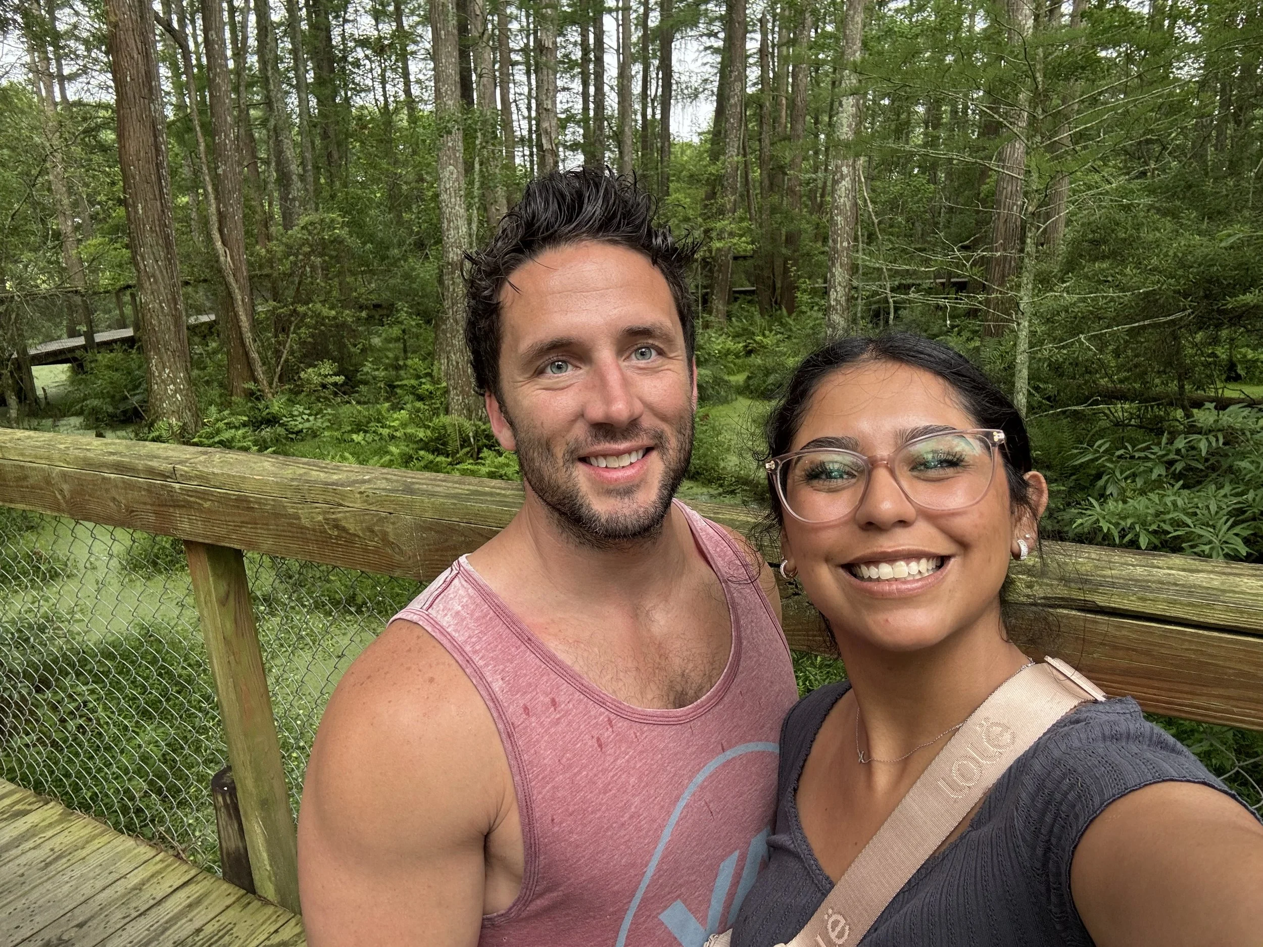 A man and woman taking a selfie outdoors in a wooded area with a wooden railing and chain-link fence in the background.