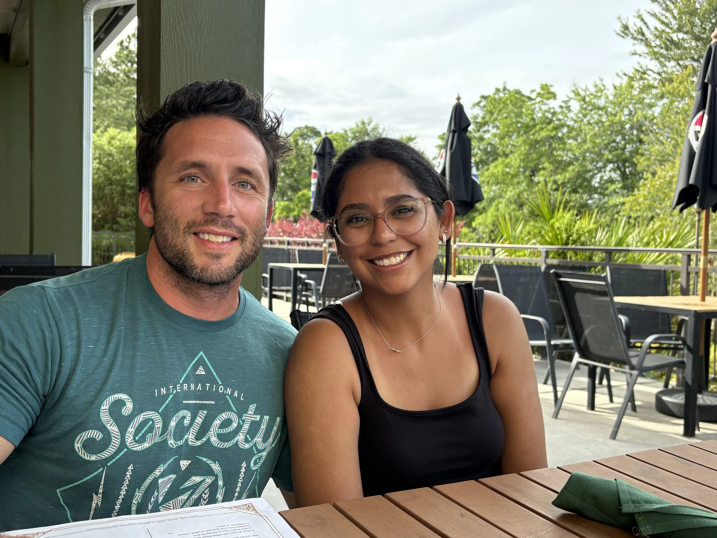 A smiling man with dark brown hair and a beard and a smiling woman with dark hair and glasses sitting at an outdoor restaurant table with a menu and a green napkin on it, with green trees and black patio umbrellas in the background.