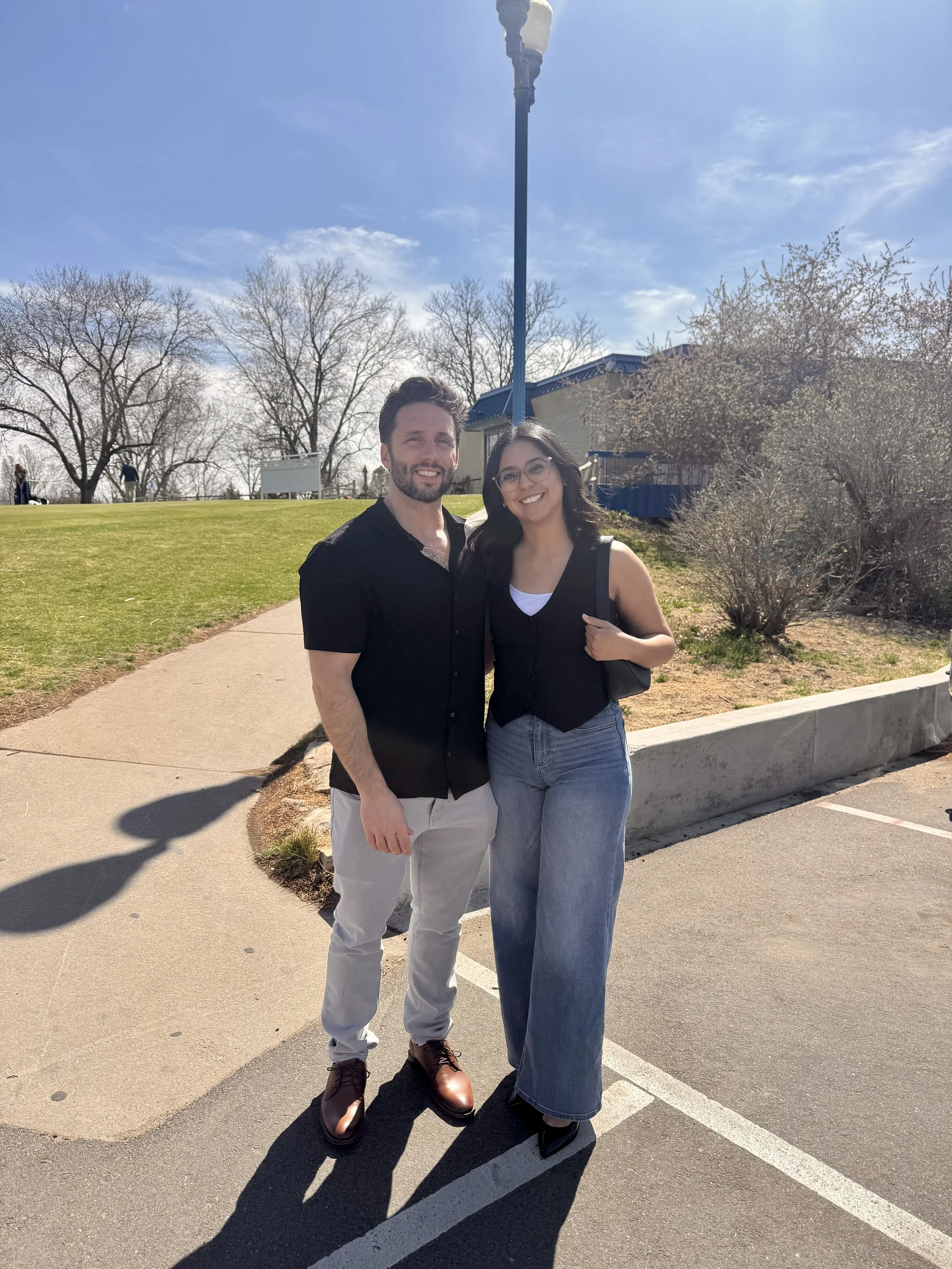 A man and woman standing together outside on a sunny day, smiling at the camera, with a grassy area, trees, and a building in the background.