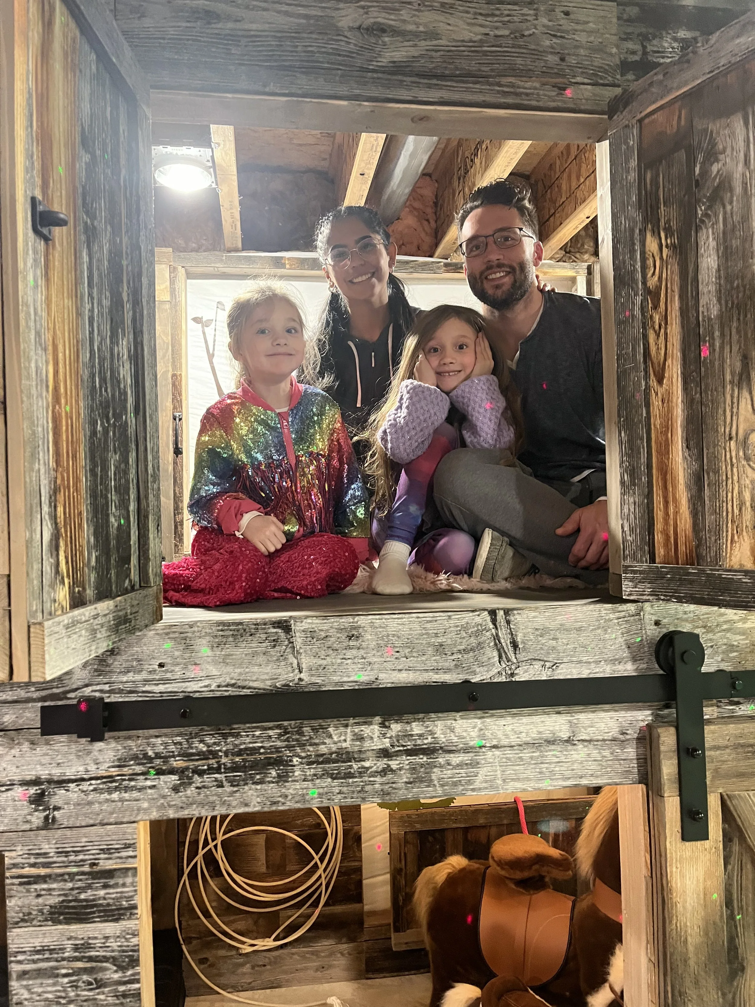 Four people, two adults and two children, are posing inside a small wooden structure. They are smiling and appear happy, with one child sitting on the floor and the other sitting on the man's lap. The setting looks cozy and rustic, with wooden walls 
