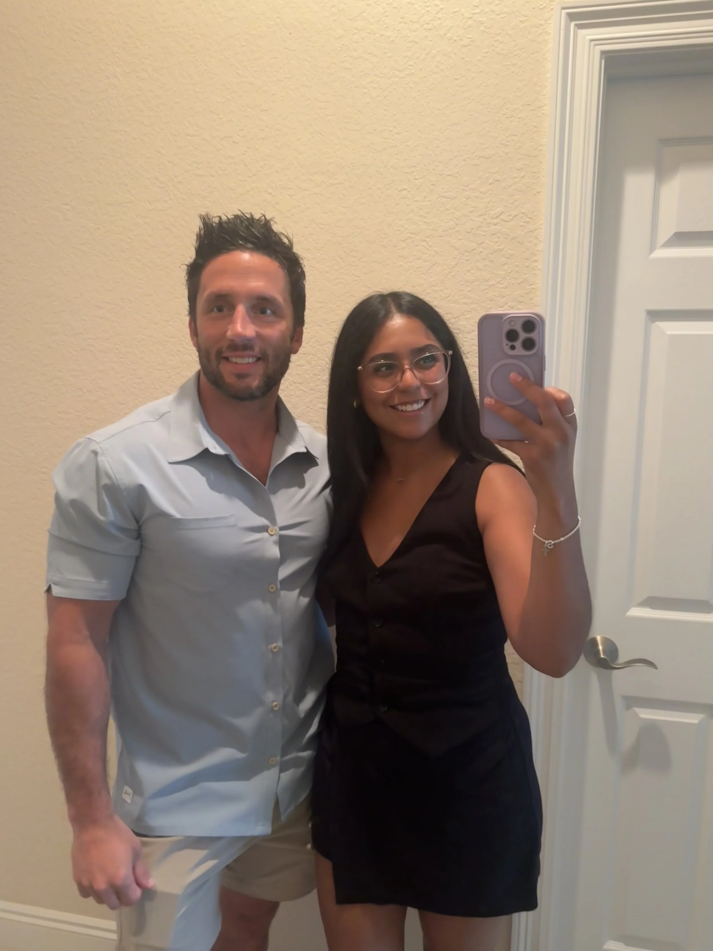A man and a woman taking a selfie in front of a mirror, both smiling. The man has dark, curly hair, a beard, and is wearing a light-colored shirt and beige shorts. The woman has long dark hair, glasses, and is wearing a black dress. They are standing