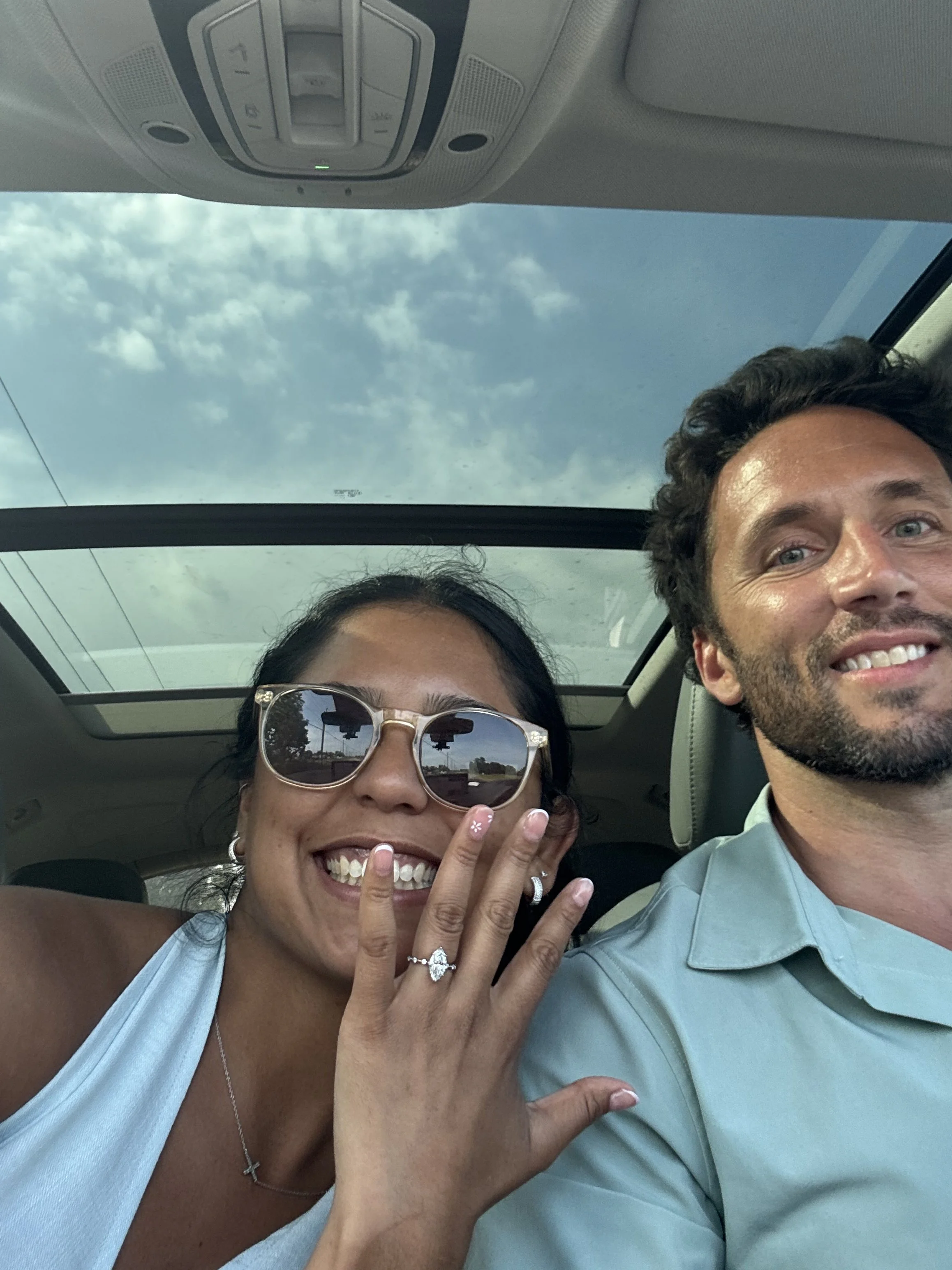 A woman with dark hair, sunglasses, and a silver cross necklace, smiling and showing her engagement ring, is sitting next to a man in a light blue shirt in a car with a panoramic sunroof, smiling at the camera.