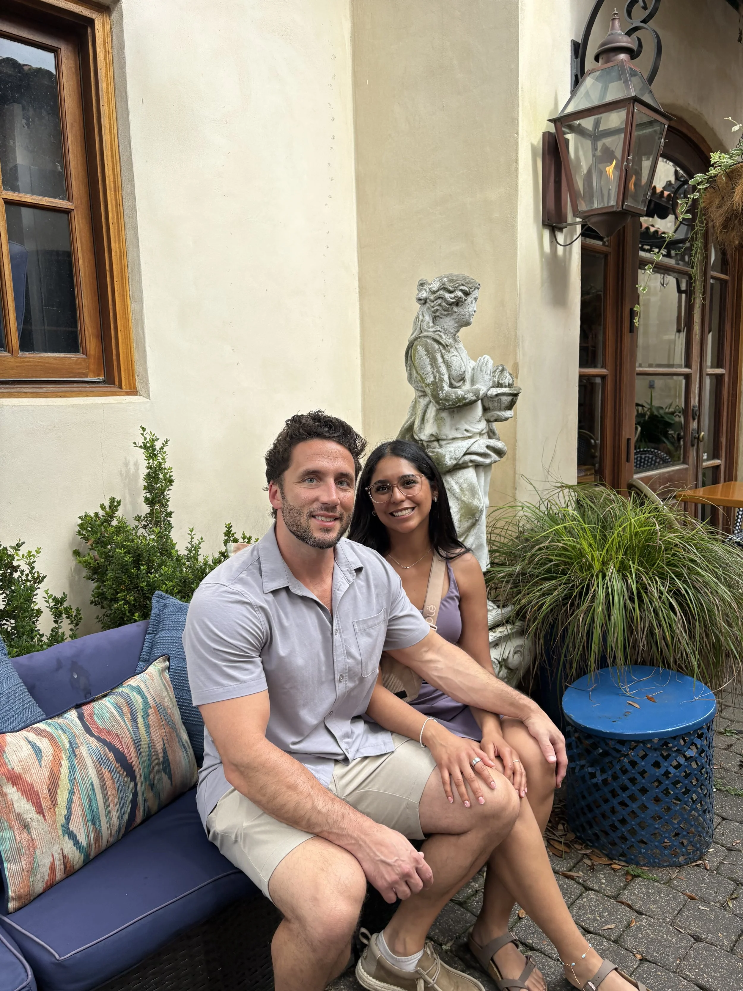 A man and a woman sitting on outdoor furniture in a garden area with plants and a stone statue of a woman holding a basket. The man has curly brown hair and beard, wearing a light gray shirt and khaki shorts. The woman has long dark hair, glasses, an