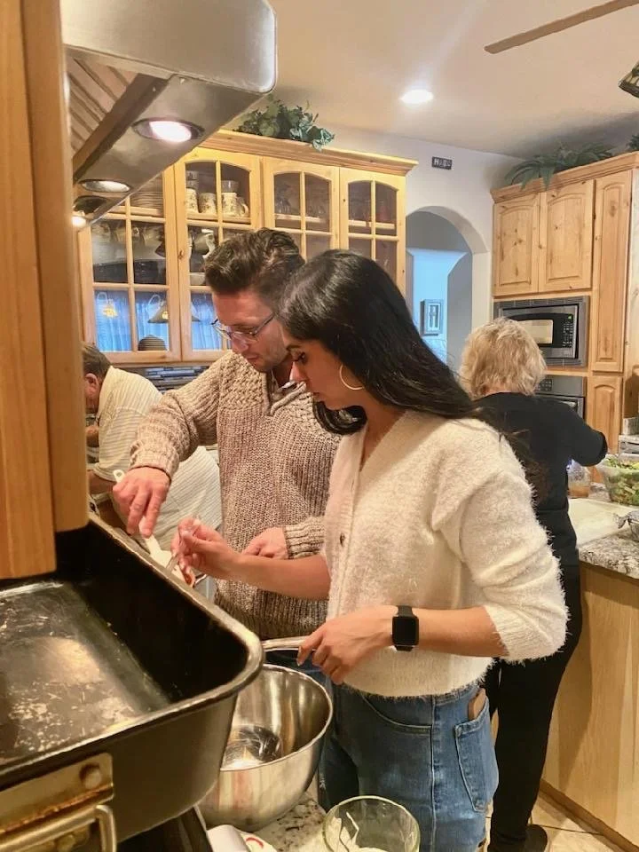 A man and woman cooking together in a kitchen, leaning over a stove and preparing food. Other people are visible in the background, and the kitchen has wooden cabinets and a microwave.