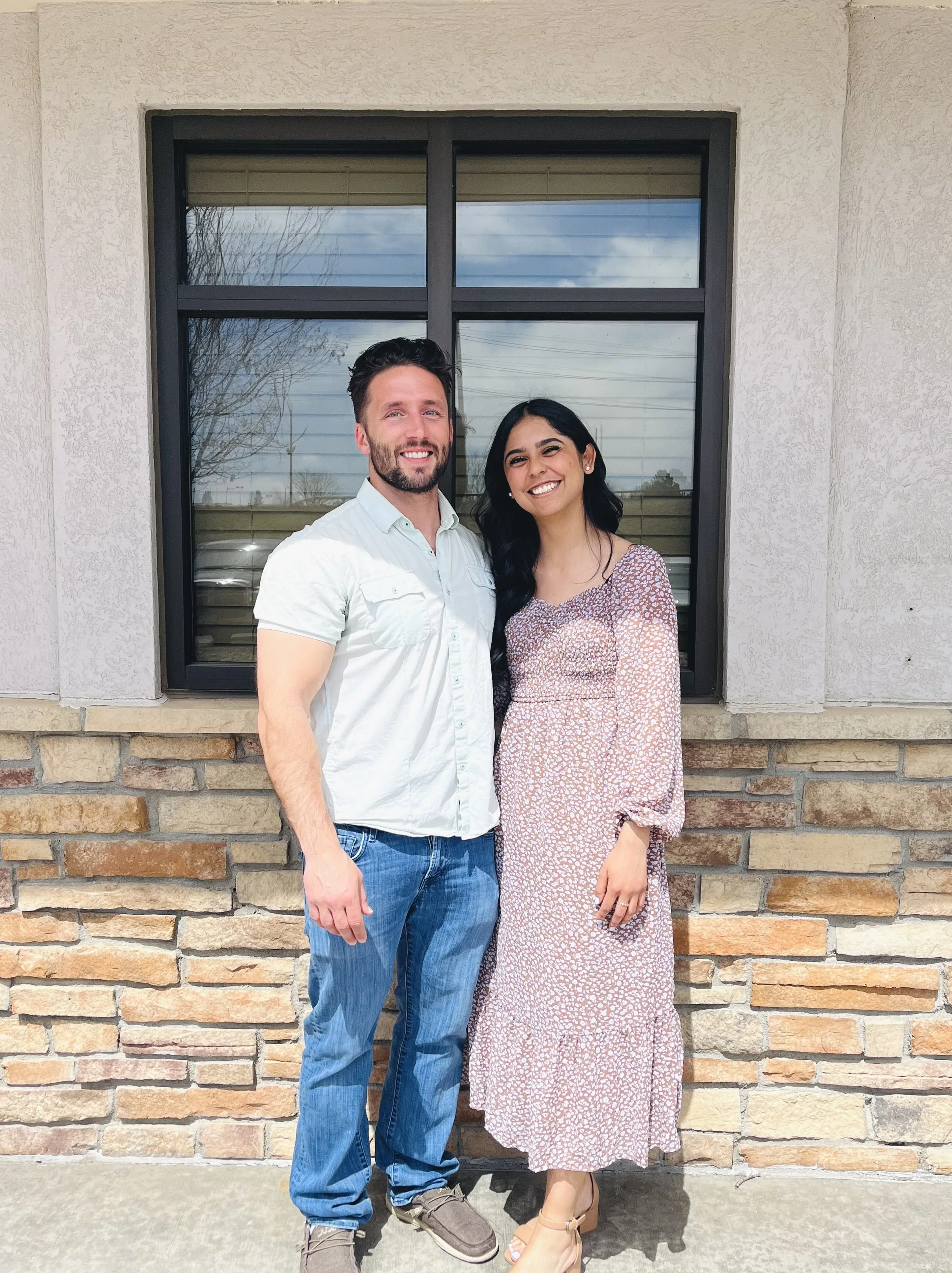 A man and a woman standing close together outside in front of a window with a stone and stucco wall behind them, smiling at the camera.