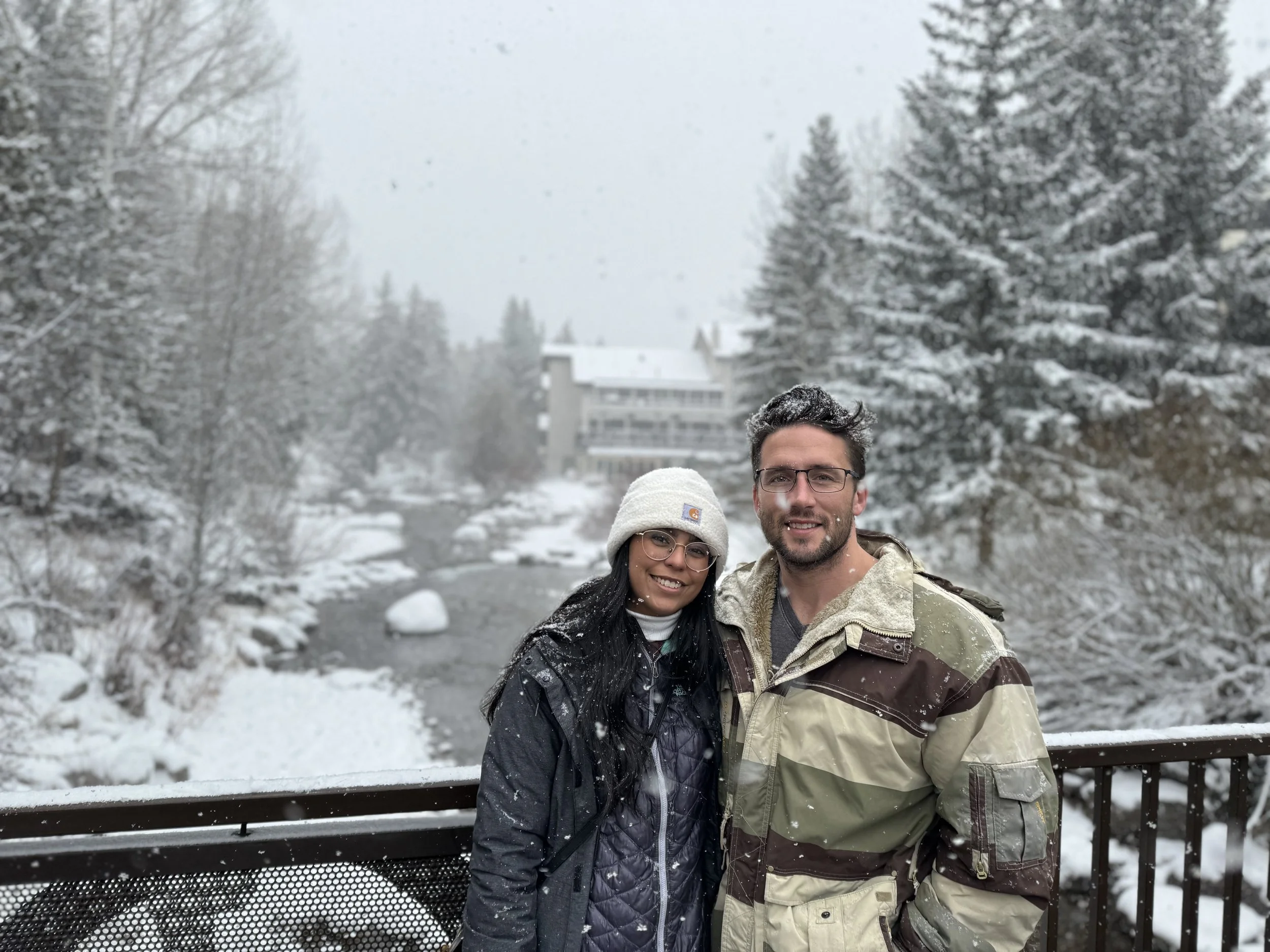A man and woman smiling and standing close together outdoors in a snowy landscape, with snow-covered trees and buildings in the background.