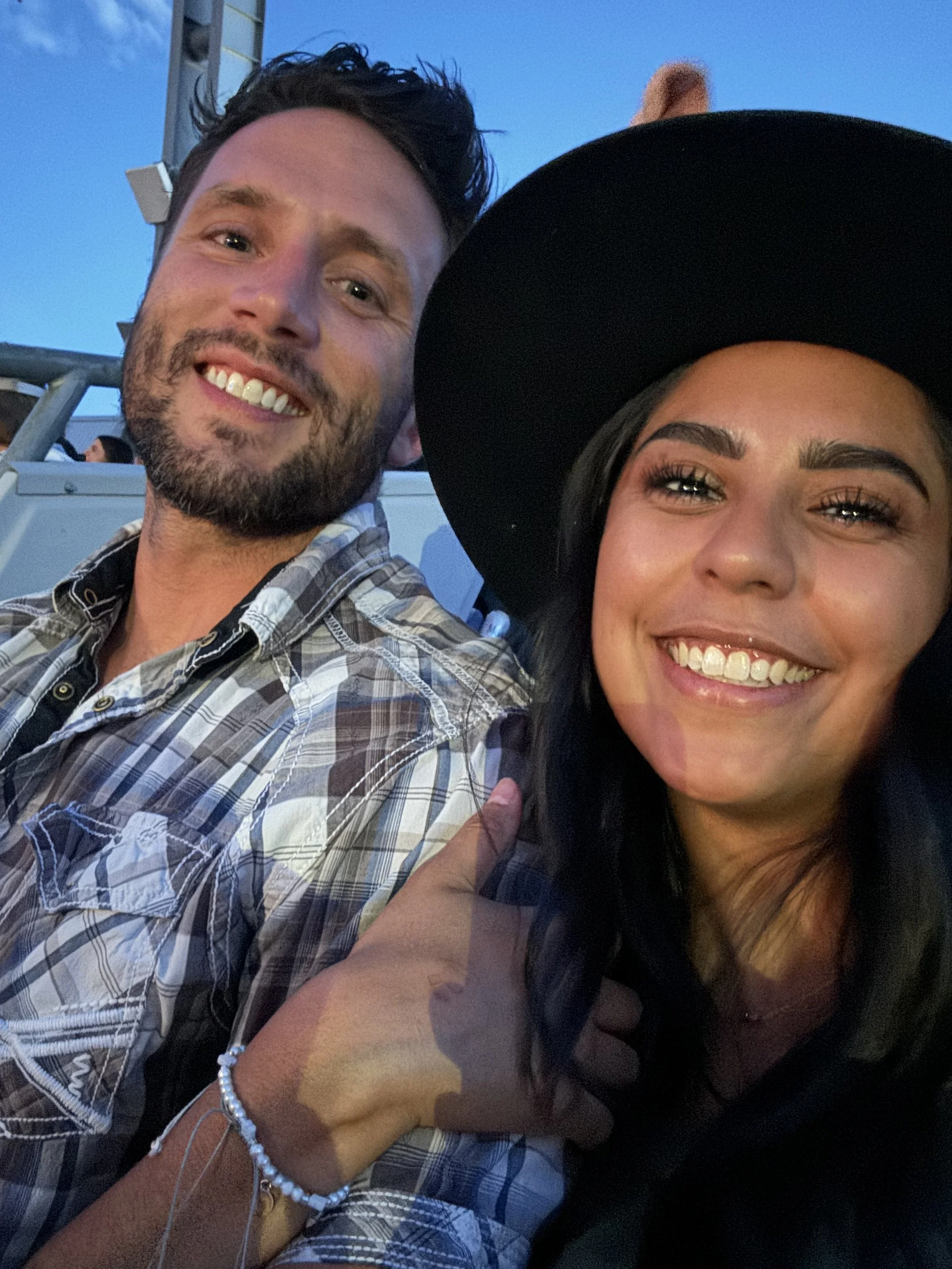 A selfie of a smiling man with dark hair and beard and a woman with long dark hair, wearing a large black hat, both smiling at the camera. The background appears to be outdoors at dusk.