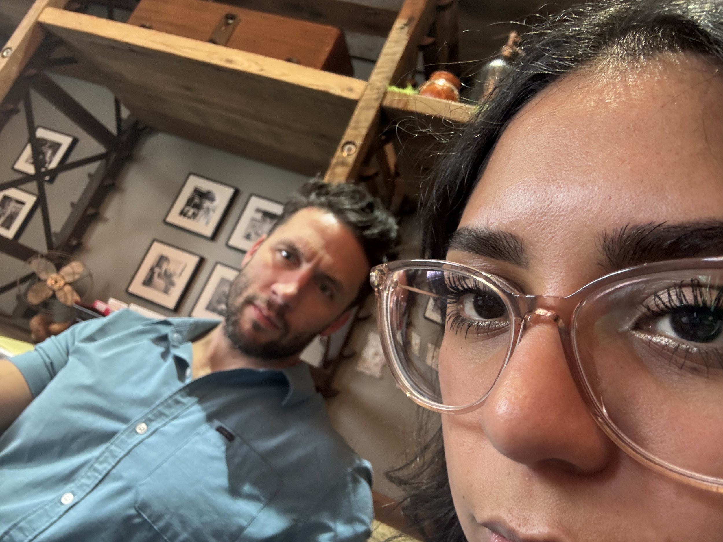Close-up of a woman's face with glasses and makeup, with a man in a blue shirt in the background, inside a restaurant or cafe with framed photos on the wall and wooden shelves.