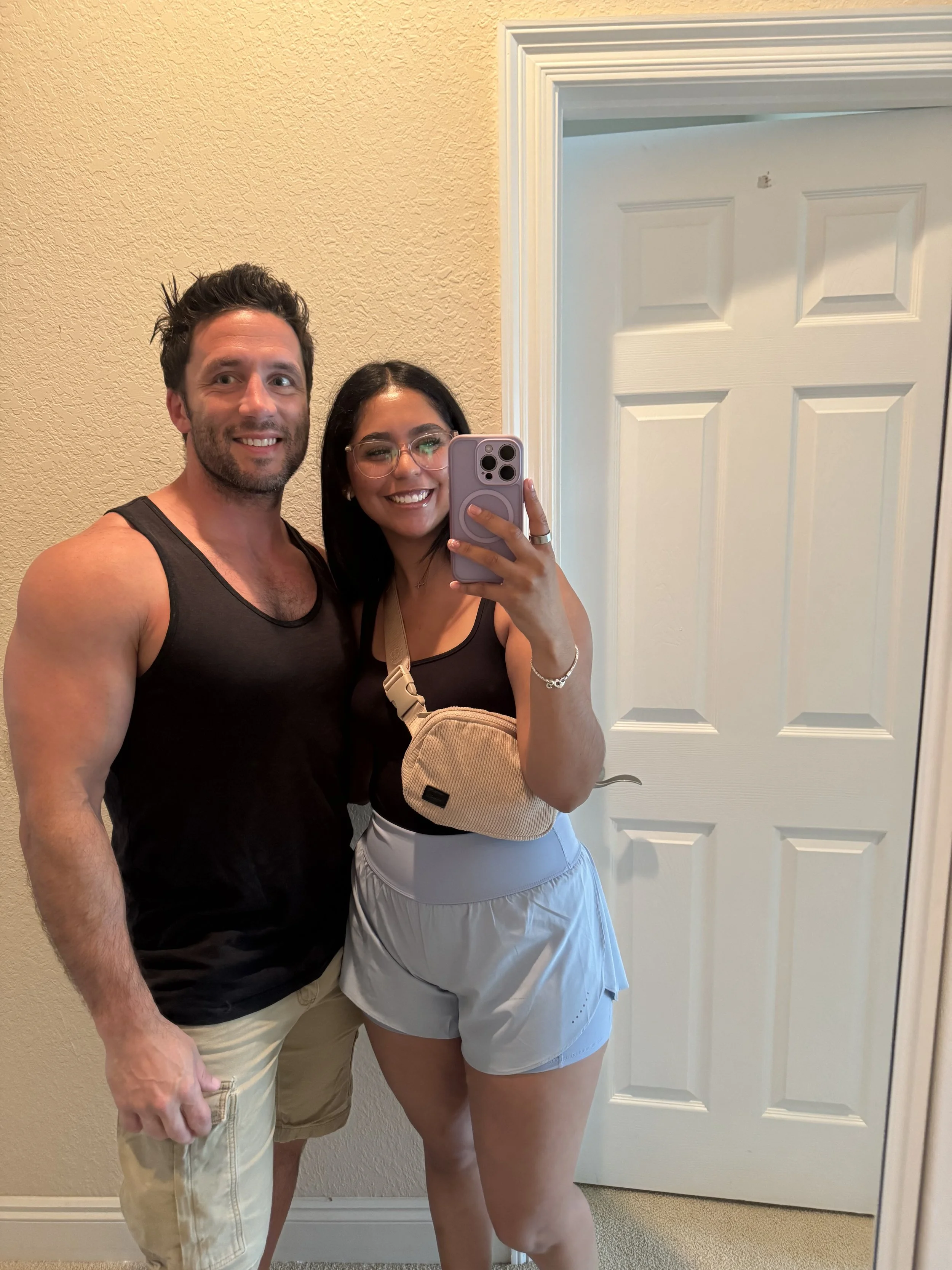 A man and a woman taking a mirror selfie indoors. The man has short dark hair, a beard, and is wearing a sleeveless black shirt and beige shorts. The woman has long dark hair, glasses, a black tank top, a white skirt, and a beige bag. She is holding 