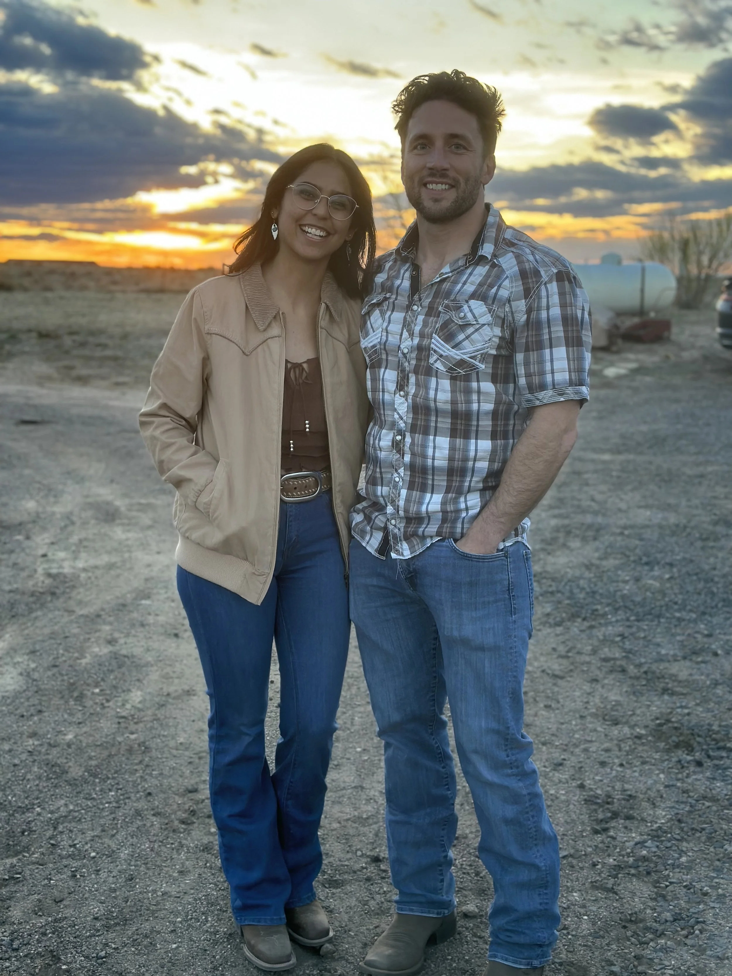 A smiling woman and man standing together outdoors during sunset, with a cloudy sky and open landscape in the background.