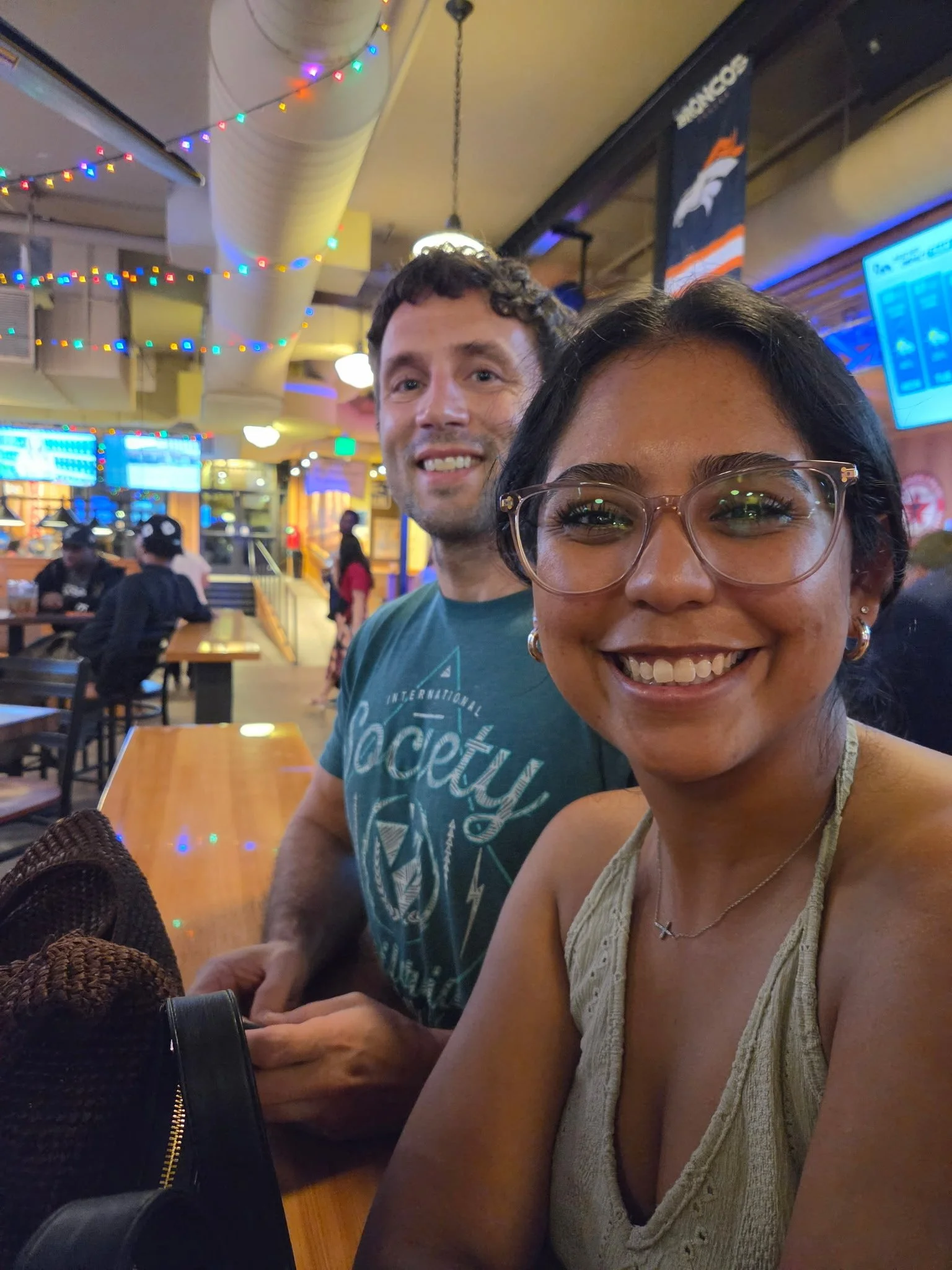 A smiling woman with glasses and a pearl necklace taking a selfie in a lively indoor restaurant or bar with colorful string lights and digital screens.