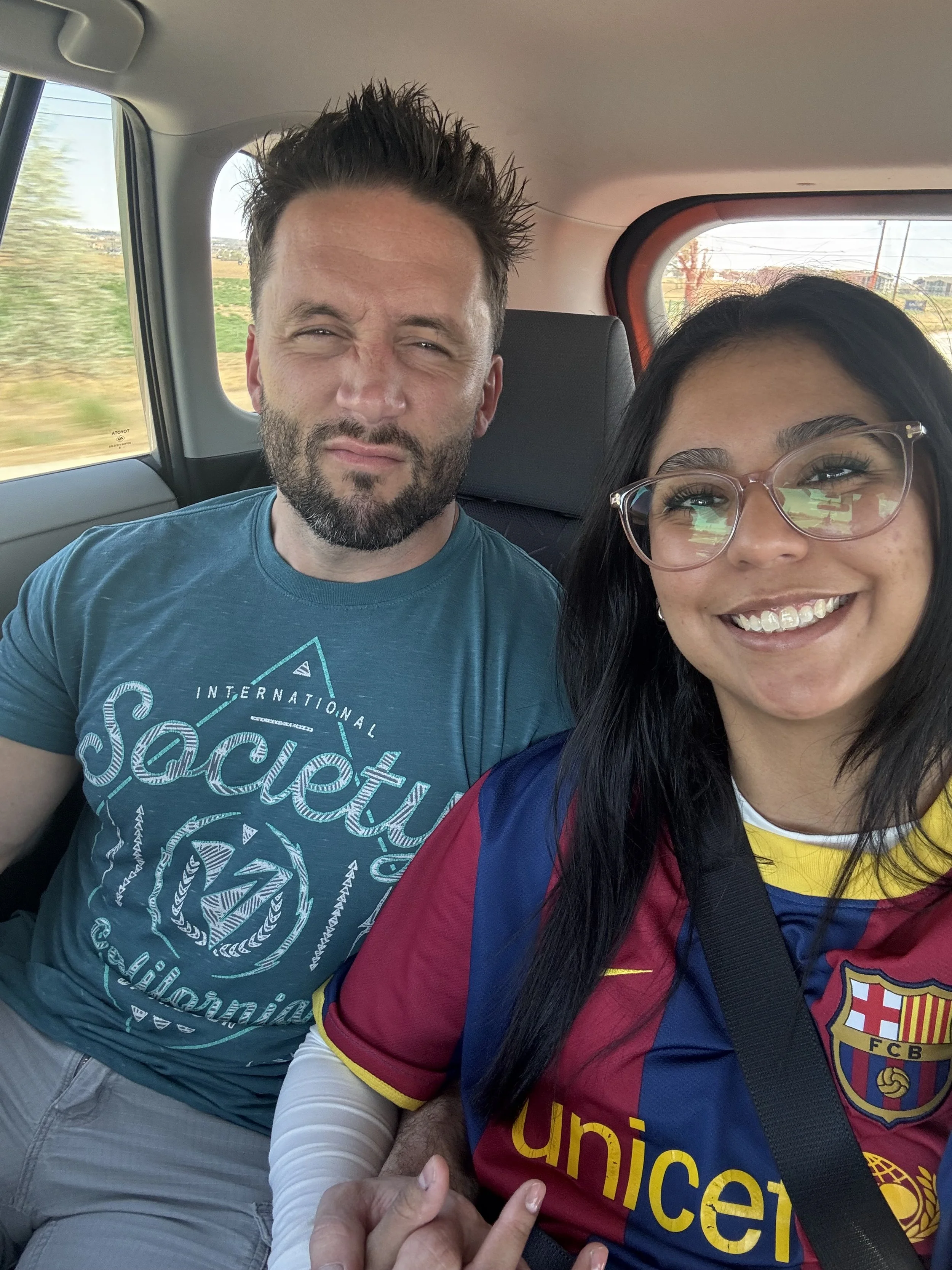 A man and a woman taking a selfie inside a car. The man has short spiked hair, a beard, and is wearing a teal t-shirt with white text. The woman has long black hair, glasses, and is wearing a colorful sports jersey with a crest on it. They are sittin