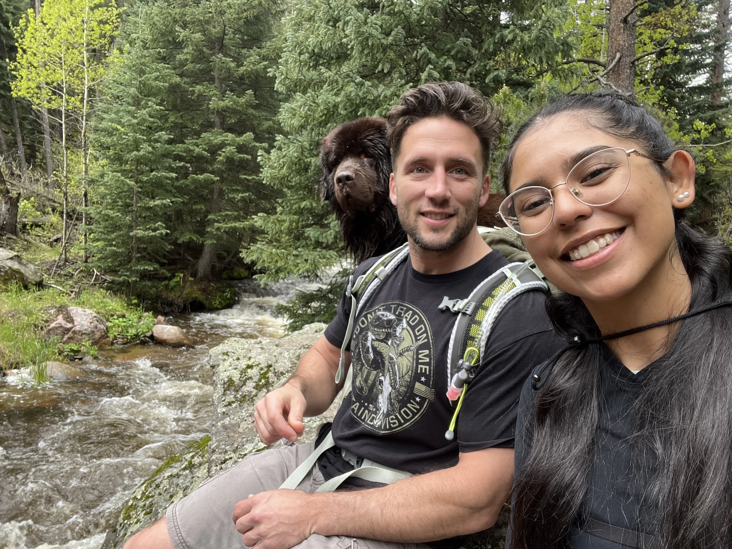 Two smiling hikers, a man and a woman, sitting by a small stream in a lush green forest. The woman has glasses and dark hair, and the man has short curly hair and a beard. A dark brown dog with wet fur is sitting behind them, partly visible.
