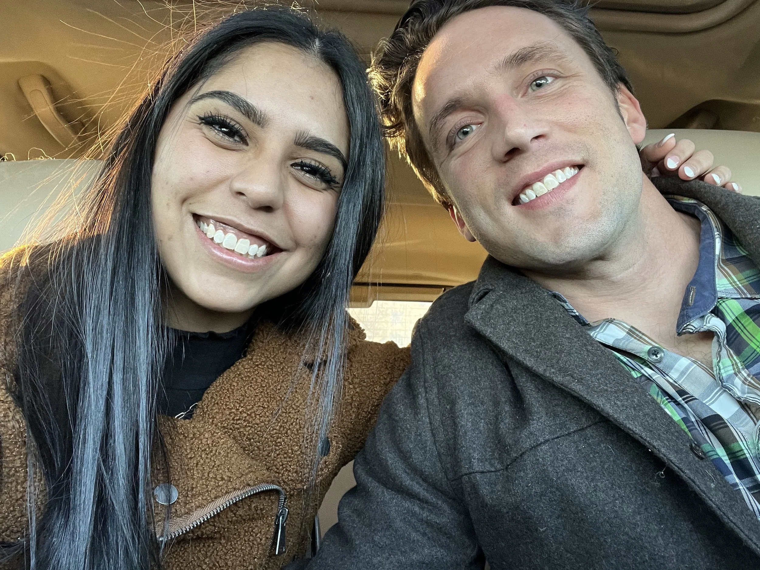 A close-up selfie of a woman with long black hair and  braces, smiling, and a man with light skin, blue eyes, dark wavy hair, smiling, both sitting inside a vehicle, with sunlight shining on their faces.