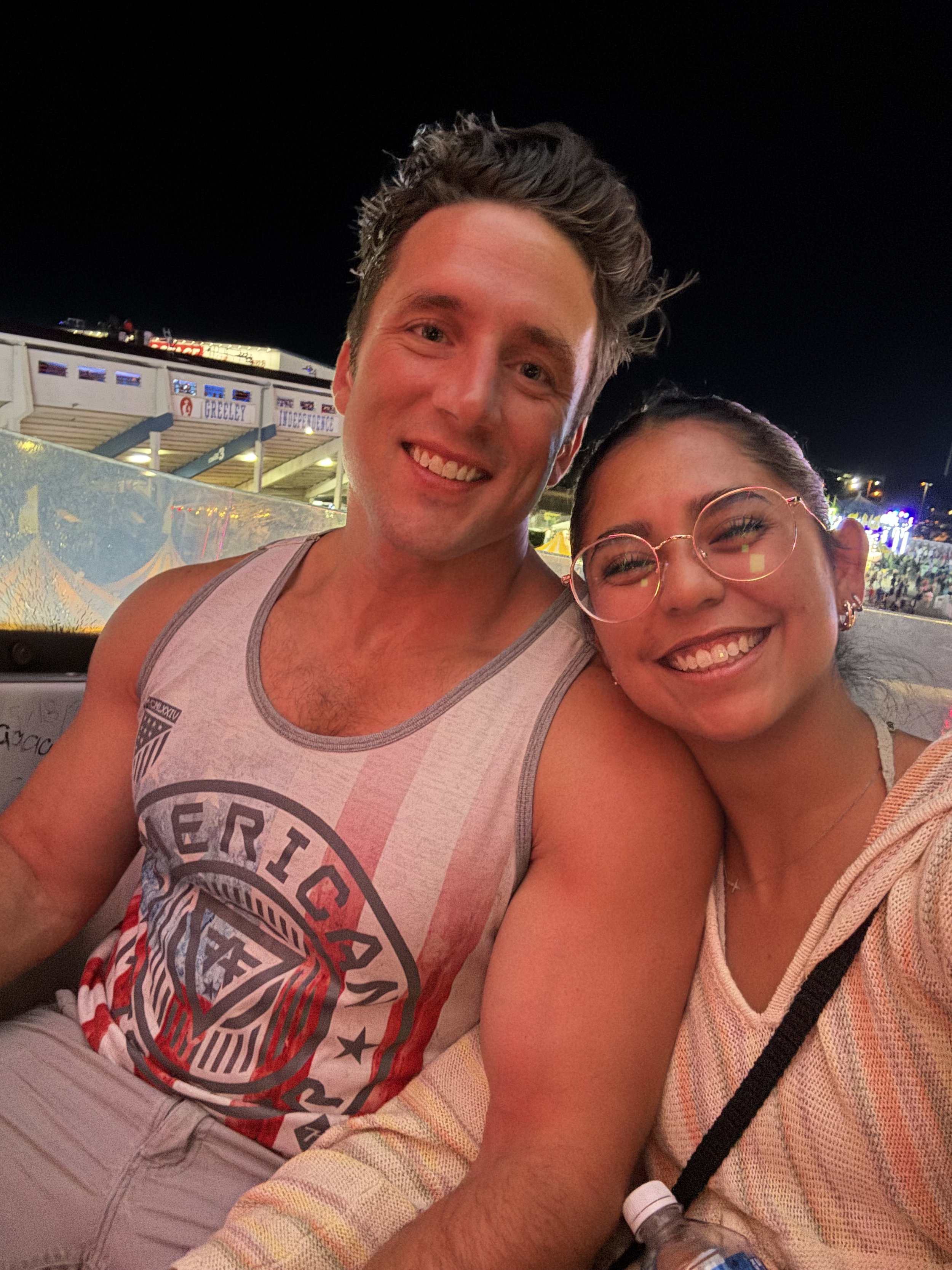 A young man and woman at night, smiling outdoors. The man has dark, styled hair and wears a sleeveless shirt. The woman has glasses and dark hair pulled back, wearing a light-colored, striped shirt or jacket. There are bright lights and a carnival or
