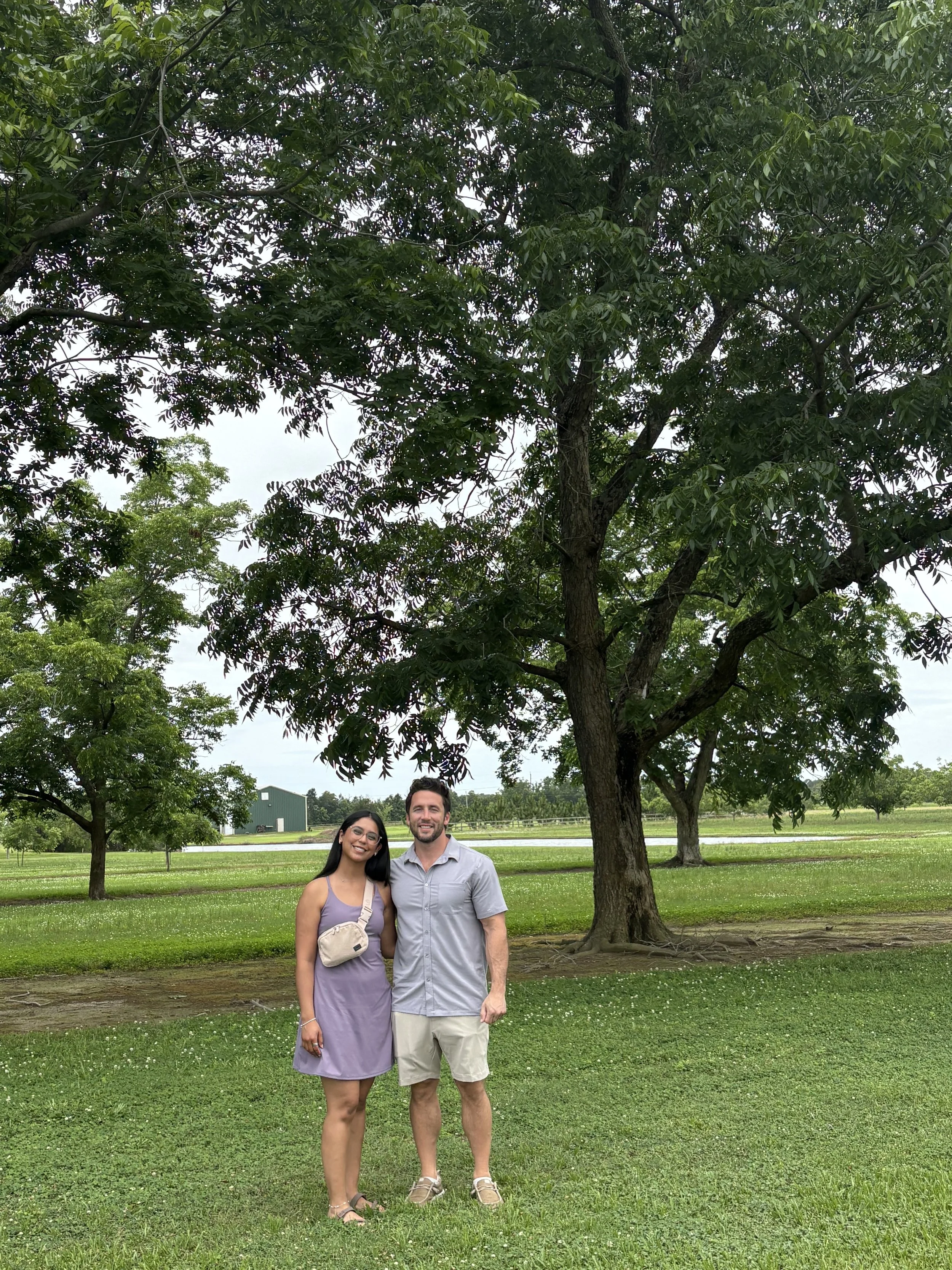 A smiling couple standing in a park with green grass and trees, near a large tree and a farm building in the background on a partly cloudy day.