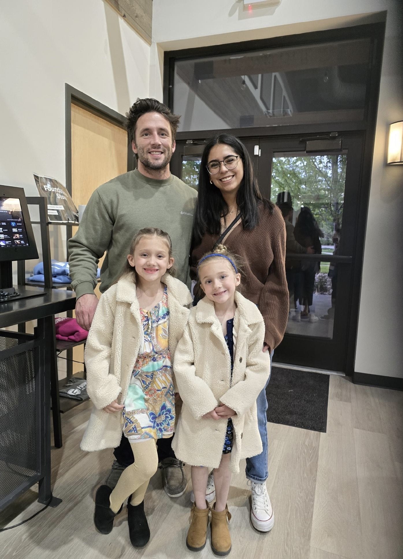 A family of four, two adults and two children, smiling and posing together inside a building near a glass door with trees visible outside. The adults are a man with dark hair and a woman with glasses, both dressed casually. The children are girls wea