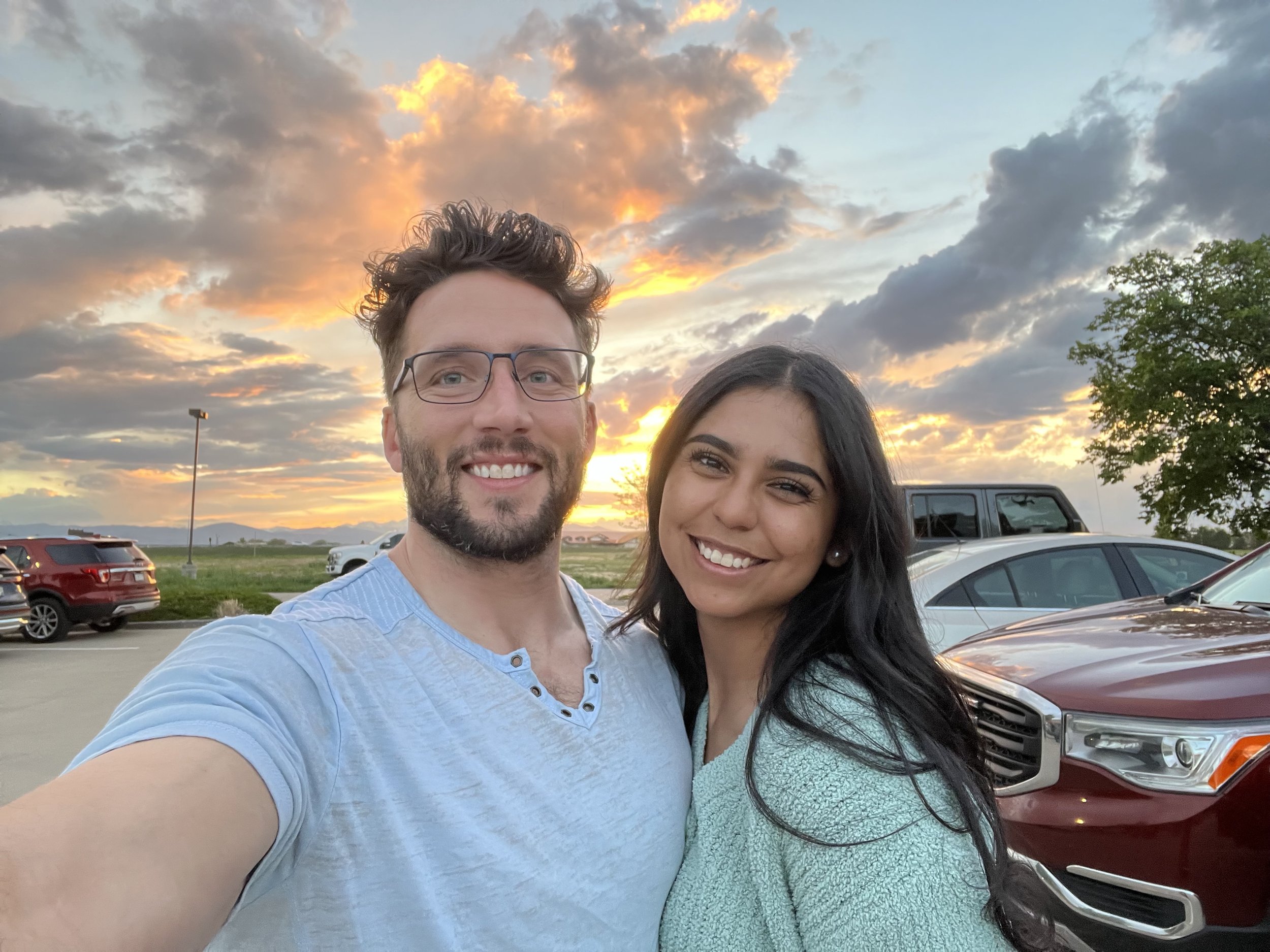 A couple taking a selfie in a parking lot during sunset with cloudy sky, cars, a tree, and mountains in the background.