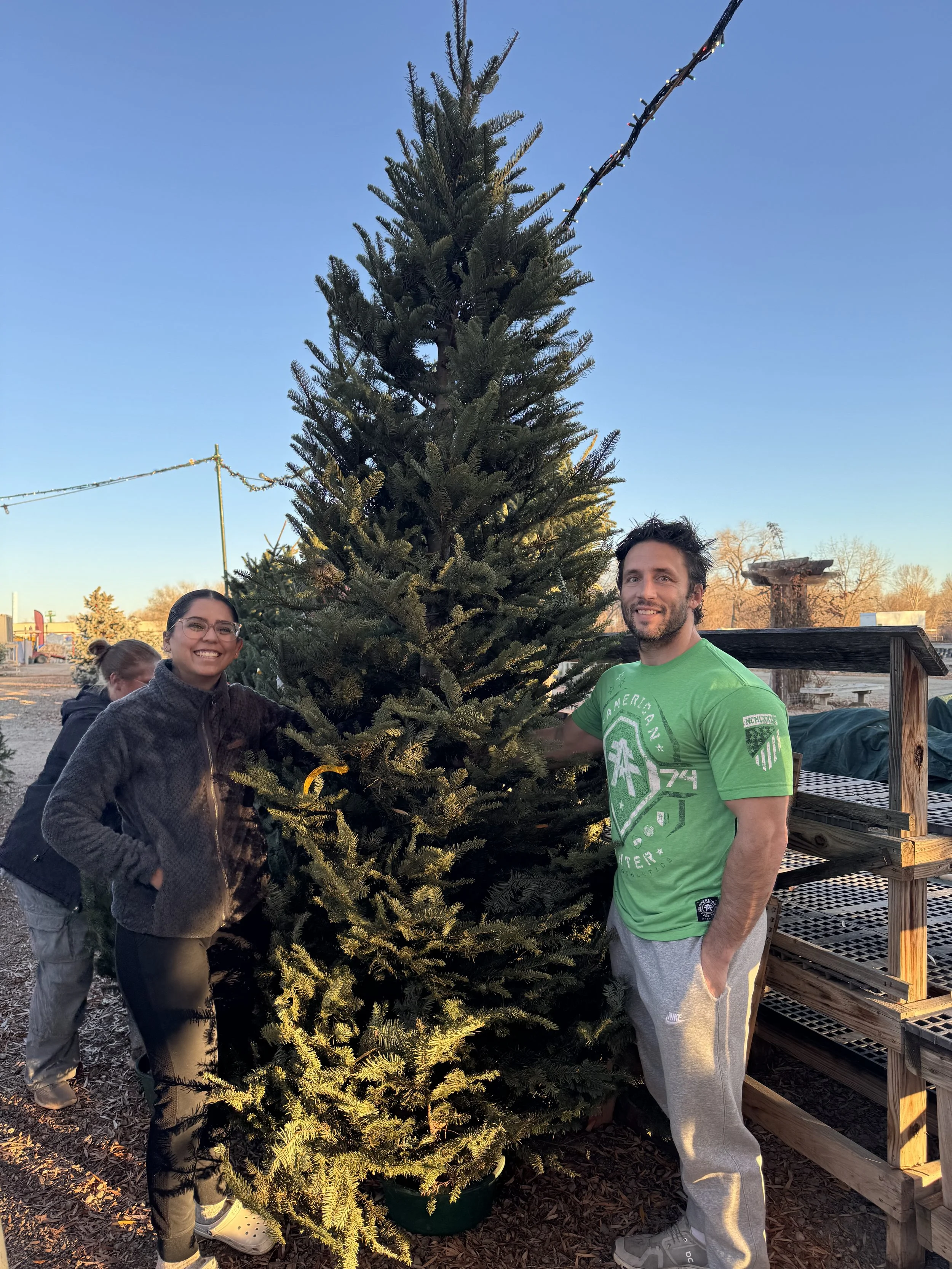 Two smiling adults, a man and a woman, standing outdoors next to a decorated Christmas tree, with a boy in the background, in a setting that appears to be a Christmas tree farm during daylight.