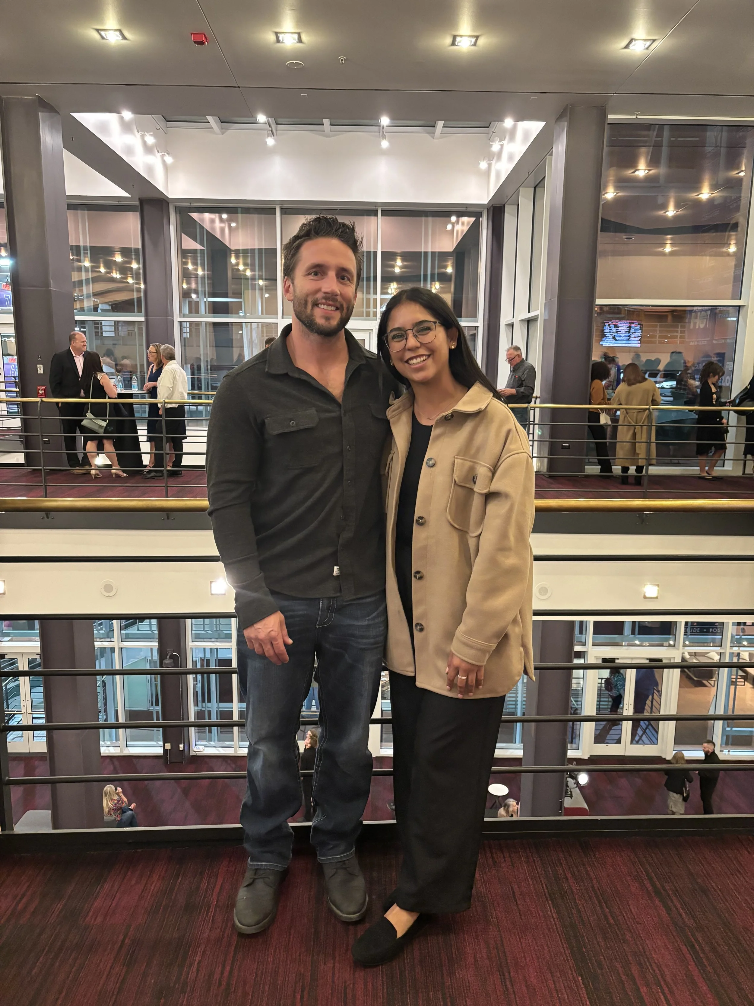 A man and woman stand close together in an indoor public space, smiling at the camera. The man has dark hair, a beard, and wears a dark jacket and jeans. The woman has long dark hair, glasses, and wears a beige jacket with black pants. Behind them, a