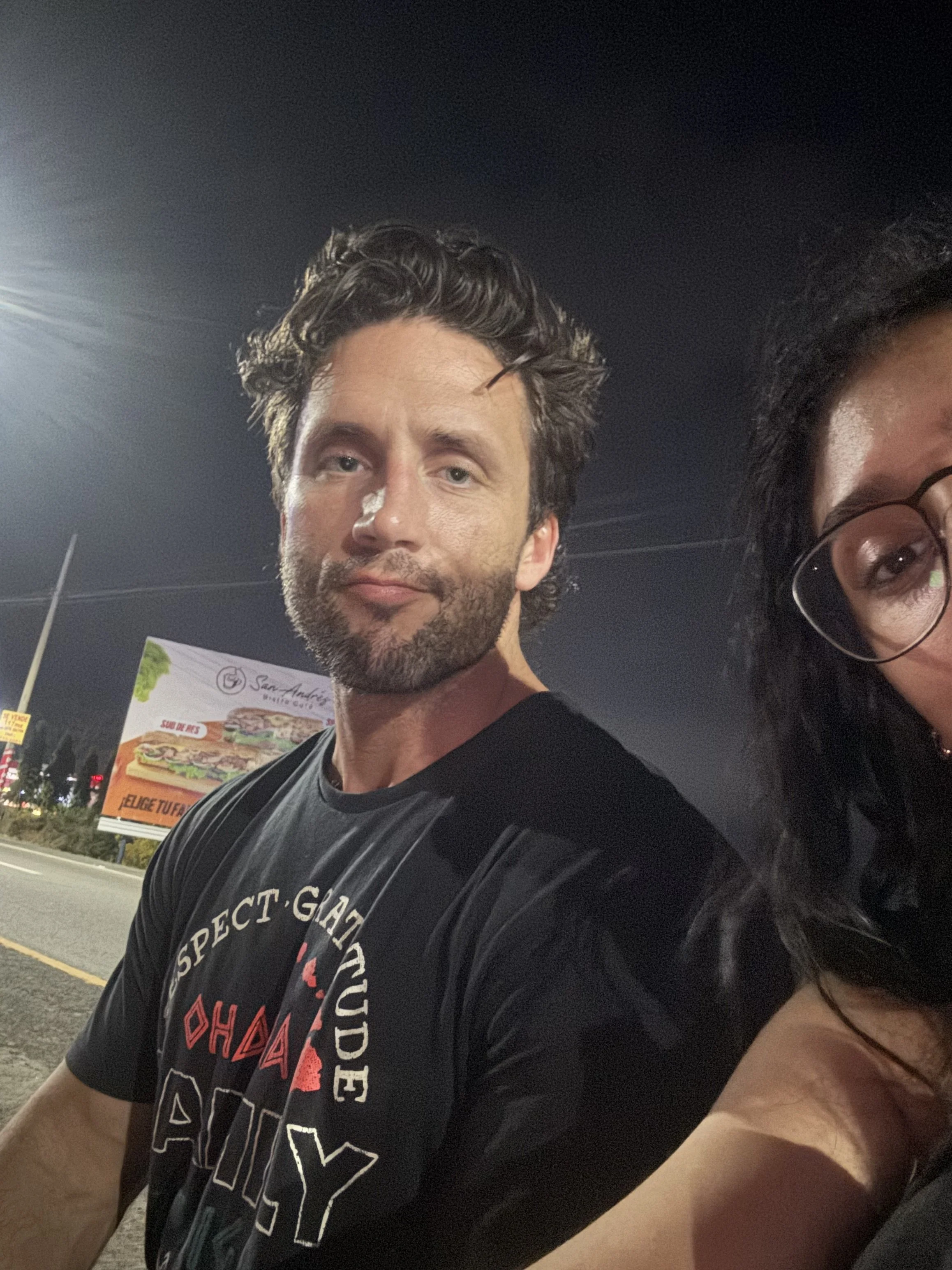 A man with dark, wavy hair, light skin, and facial hair taking a selfie at night. Part of a woman with glasses and dark hair is visible on the right. In the background, there are billboards, including one with a sandwich image and a street sign. The 