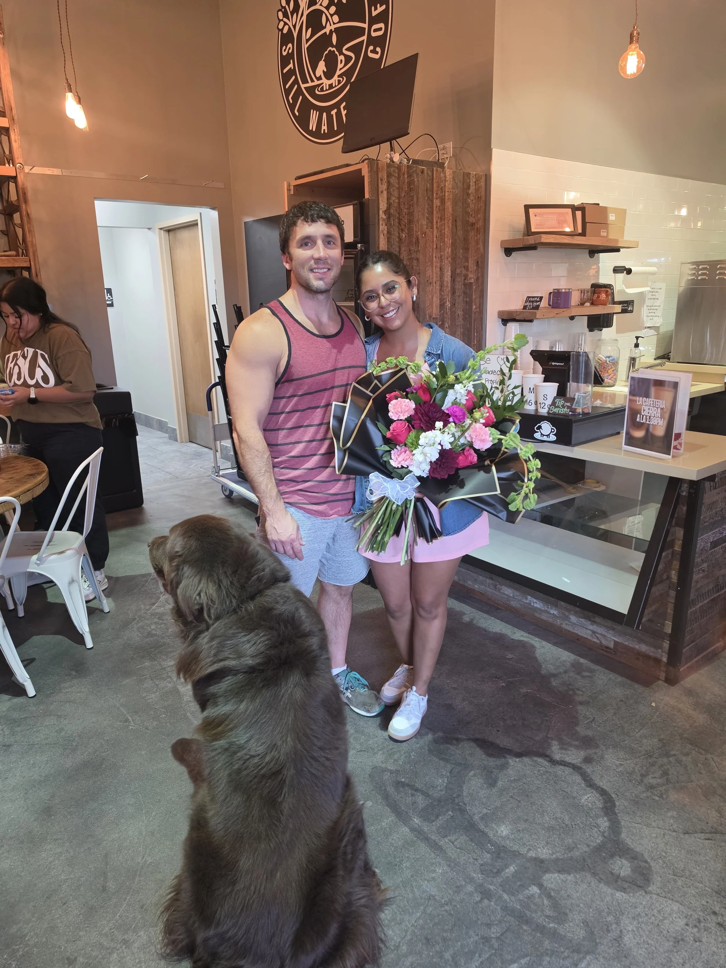A man and woman smiling at the camera, holding a bouquet of flowers, with a large dog in the foreground inside a coffee shop.