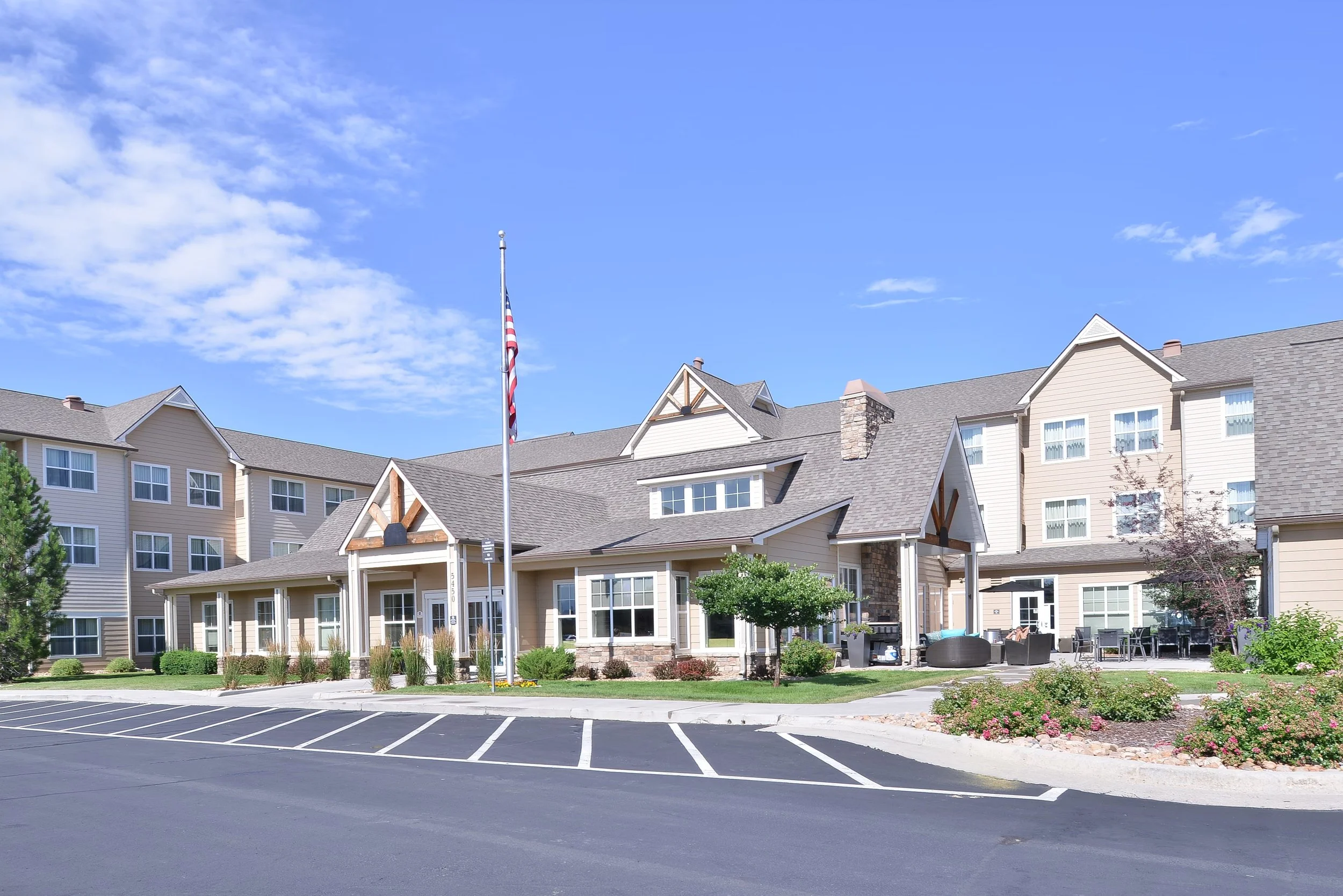 A residential building complex with beige and cream-colored houses, a parking lot with white-lined spaces, a flagpole with an American flag, landscaped greenery, and an outdoor seating area under a blue sky with some clouds.