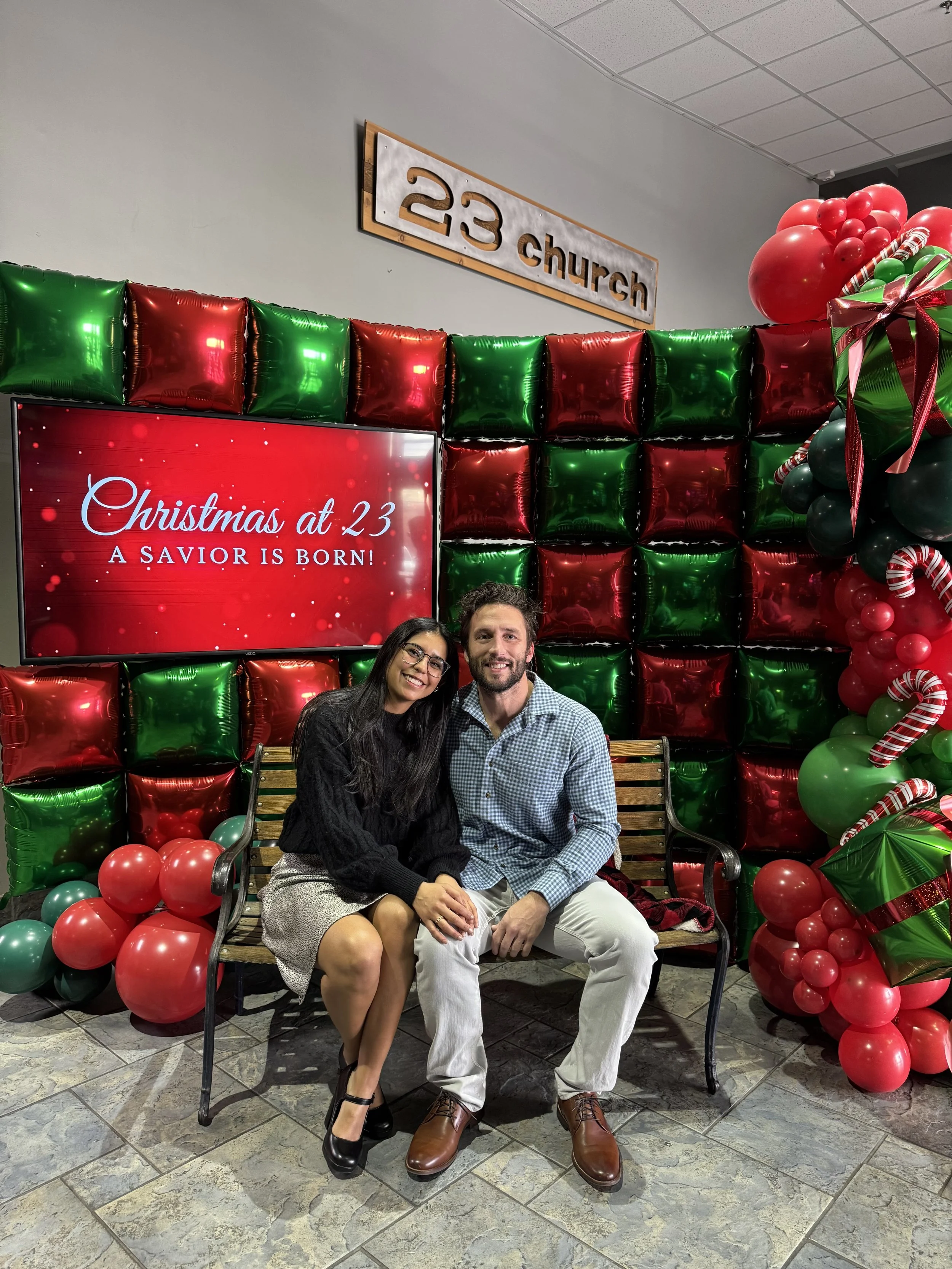A man and a woman sitting on a wooden bench at a Christmas celebration. They are smiling, with the woman resting her hand on the man's knee. Behind them is a festive backdrop with green and red balloons and a sign that reads "Christmas at 23, A Savio