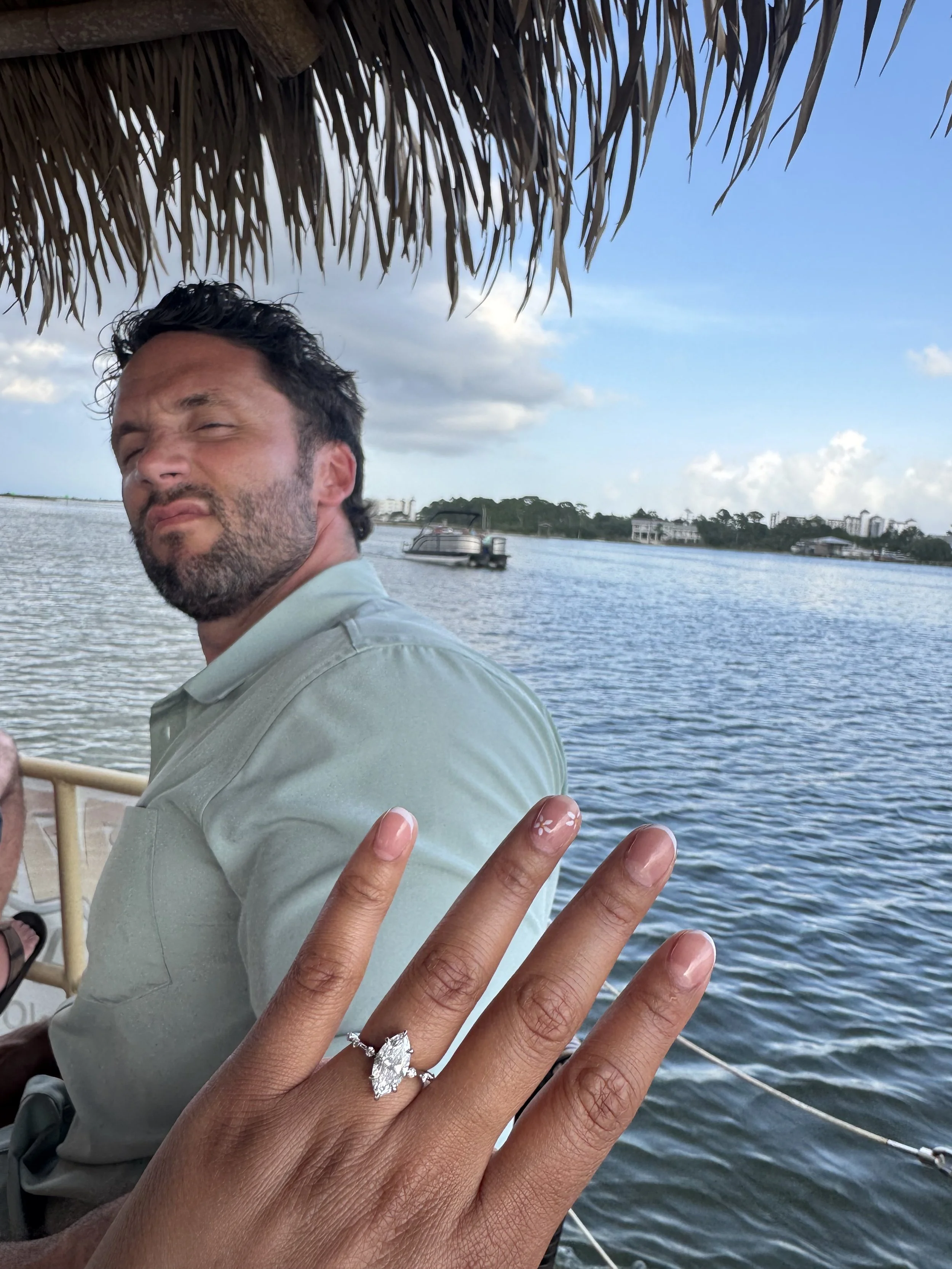 A man with dark hair and a beard on a boat, with a hand in the foreground showing a diamond ring. The background shows water, a boat, and a distant shoreline with trees and buildings, under a partly cloudy sky.