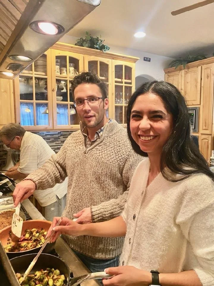 A smiling woman and a man cooking Brussels sprouts in a kitchen with glass cabinet doors and wooden cabinets.