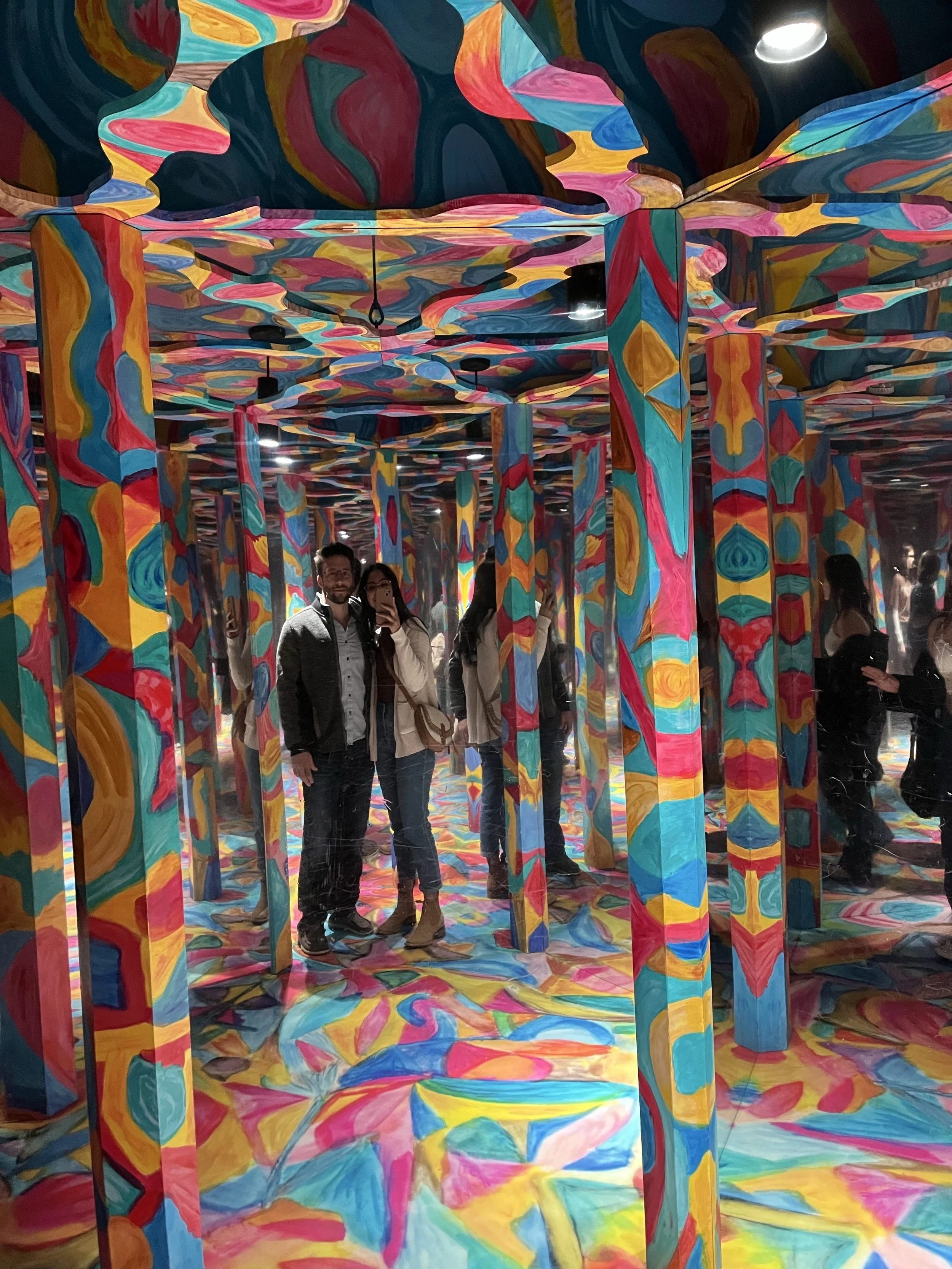 People posing inside a mirrored room with vibrant, swirling, colorful patterns on the walls, ceiling, and floor.