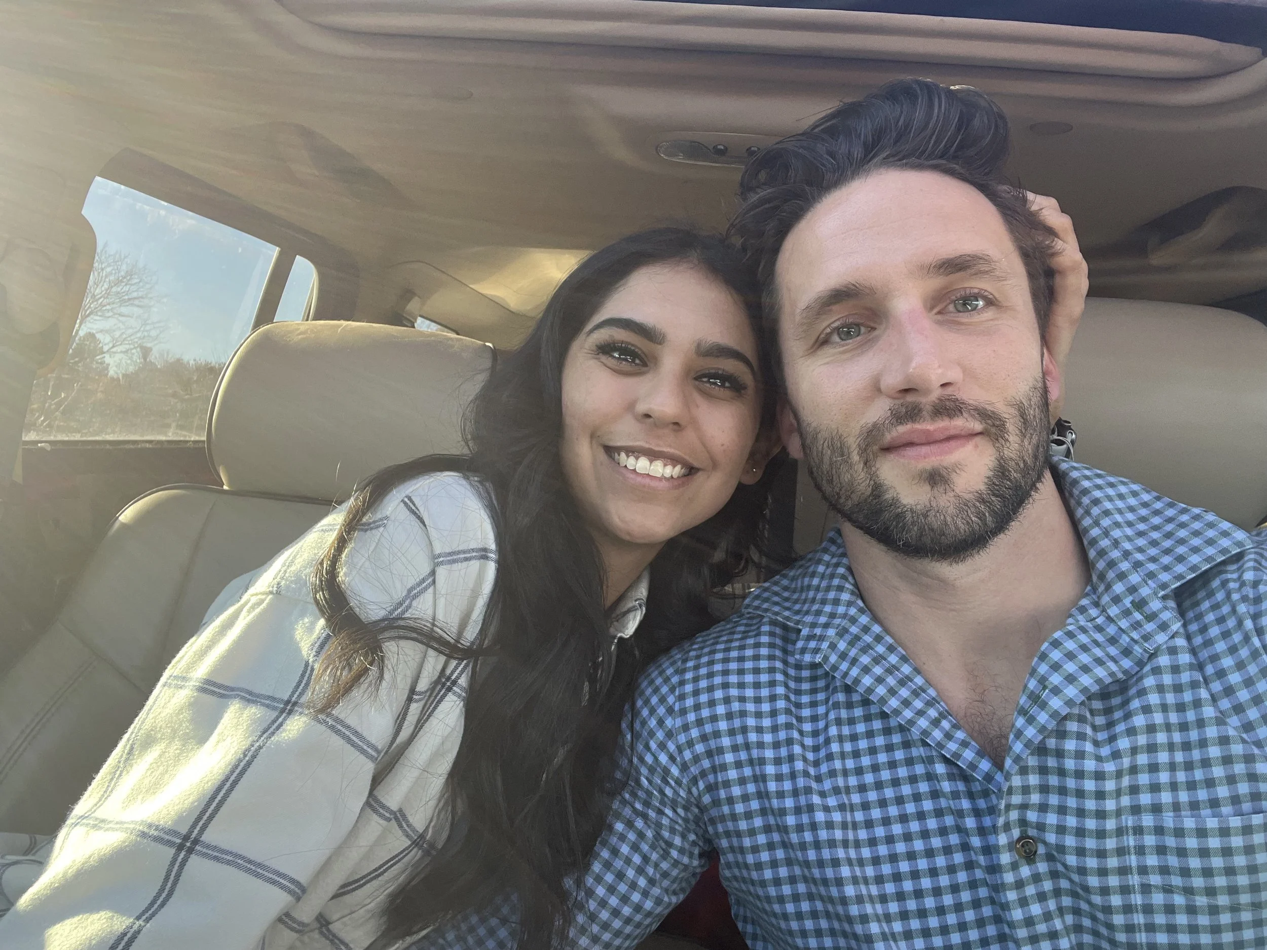 Close-up of a smiling woman with dark hair and a man with light hair and a beard, sitting together in a vehicle. The woman is wearing a plaid shirt and has earrings, while the man is wearing a blue checkered shirt. They are taking a selfie, with sunl