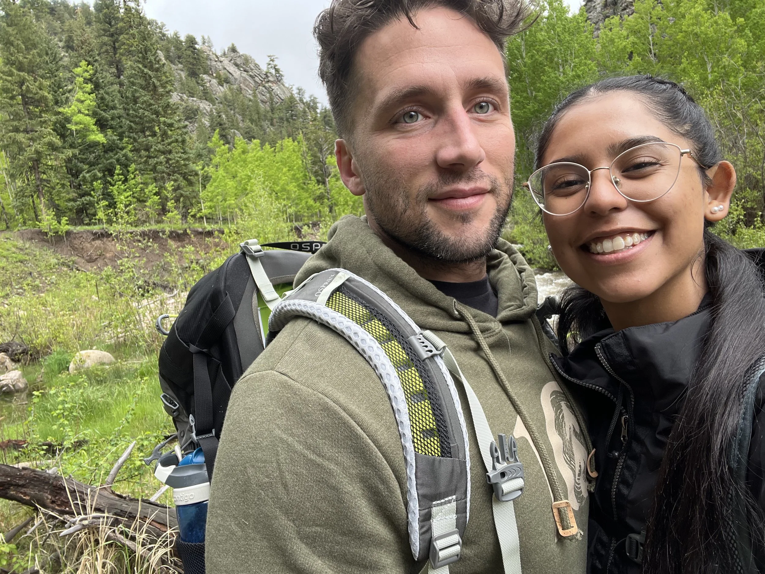 A man and a young girl smiling together outdoors in a wooded area, with trees, rocks, and a mountain in the background.