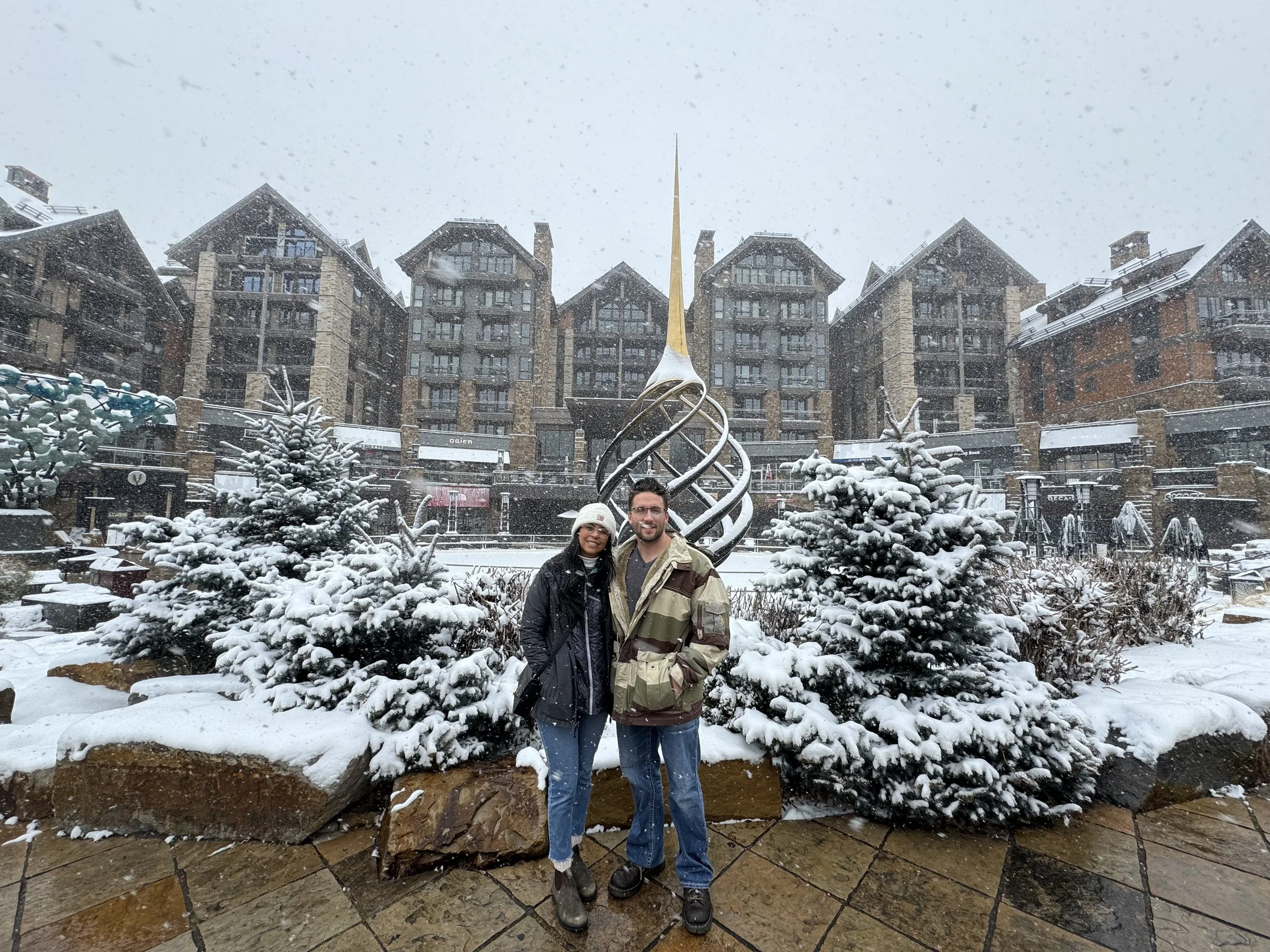 A couple stands close together outdoors on a snowy day, smiling at the camera. They are in front of snow-covered bushes, with multi-story buildings and a mountain landscape in the background.