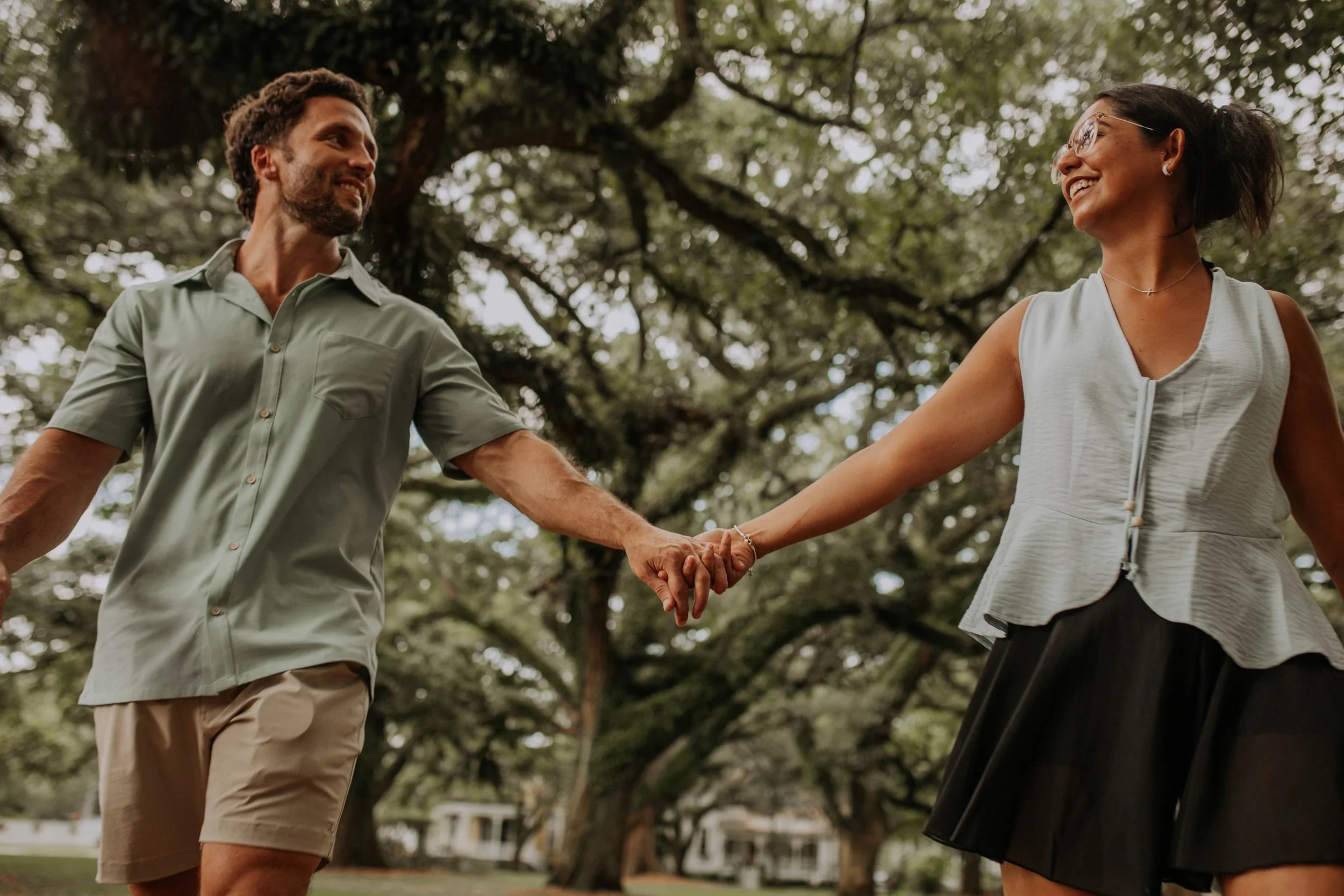 A man and a woman holding hands and smiling at each other outdoors under a large tree.