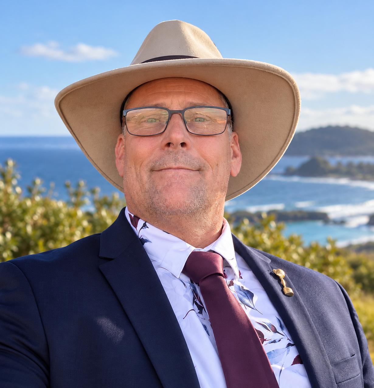 A man wearing glasses, a wide-brimmed hat, a navy blazer, a patterned shirt, and a maroon tie, standing outdoors near the coast with ocean, hills, and blue sky in the background.