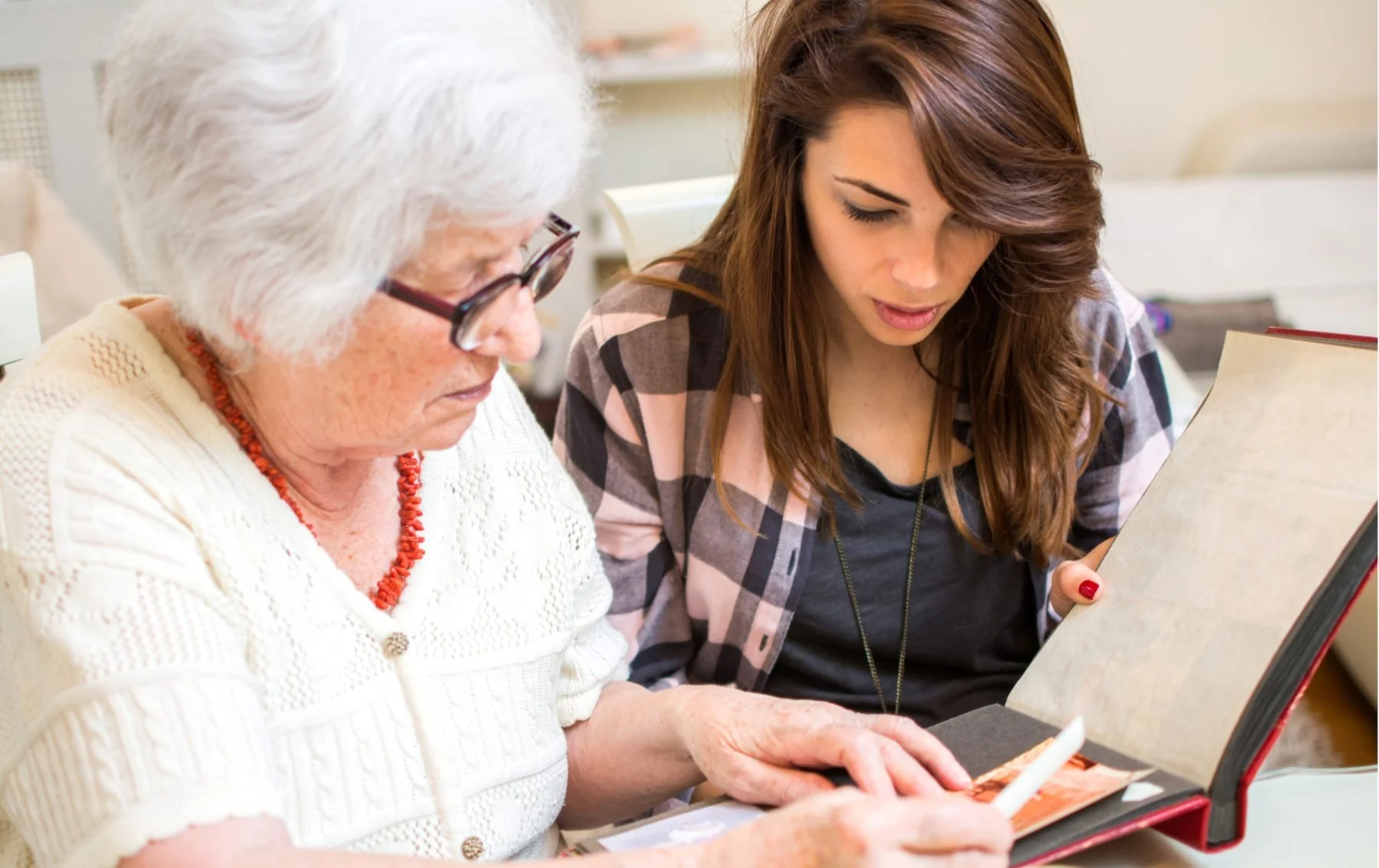 An elderly woman with white hair and glasses looking at a photo album with a young woman with brown hair and a plaid shirt.