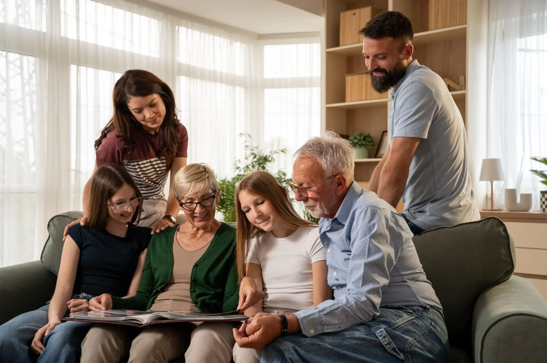 Family gathered on a sofa, looking at a photo album together, in a well-lit living room with large windows and bookshelves.