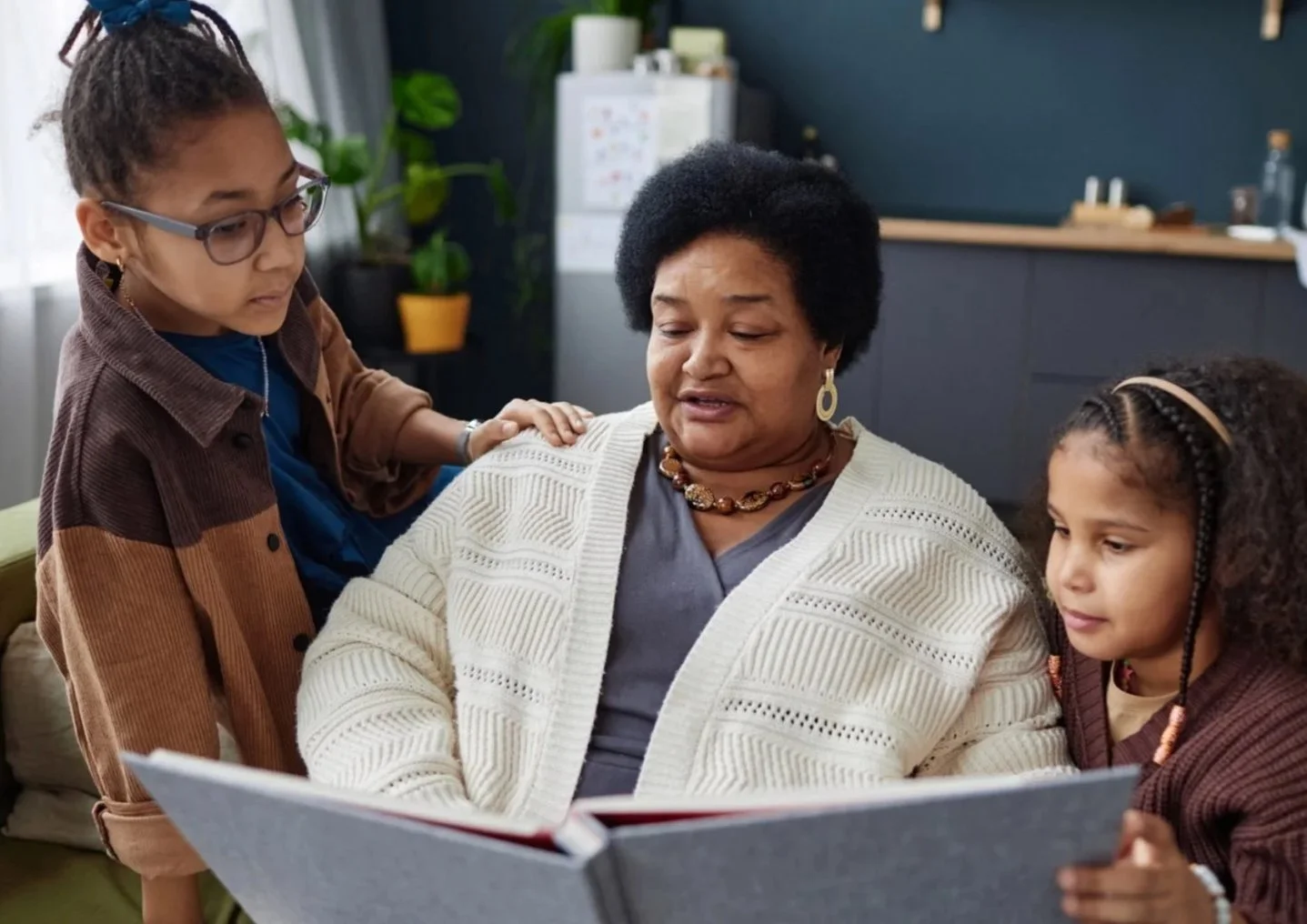 A woman with short black hair and earrings is sitting on a couch, reading a book to two young girls in a cozy, modern room with plants and kitchen in the background.