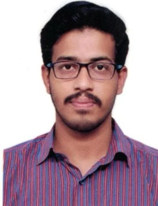 A young man with black hair, glasses, and a mustache, wearing a striped button-up shirt, standing against a plain white background.