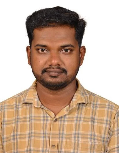 A man with short black hair, a beard, and brown skin, wearing a yellow plaid button-up shirt, looking directly at the camera against a plain white background.