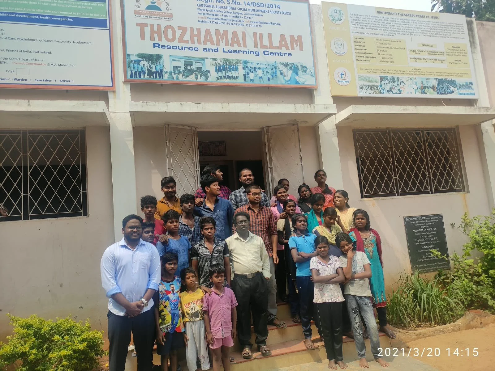 A group of people, including children and adults, standing outside a building with informational posters on the wall, some of whom are smiling at the camera.