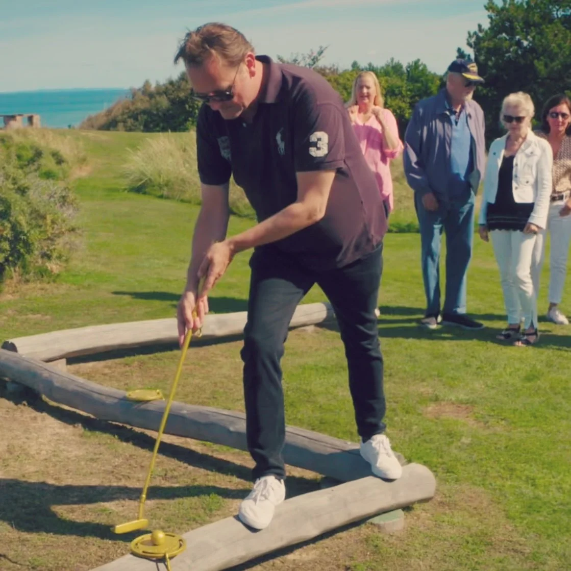En mand spiller minigolf på en udendørs bane, mens en gruppe mennesker ser til i baggrunden.