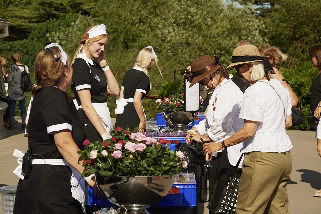 Kvindelige tjenere i sort uniform, der serverer mad til gruppe af ældre kvinder med solhatte ved udendørs buffet.