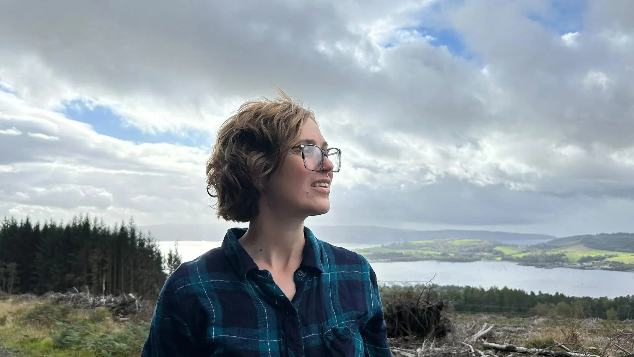 A young woman with short wavy hair and glasses stands outdoors in front of a landscape with trees, water, and green hills under a cloudy sky.