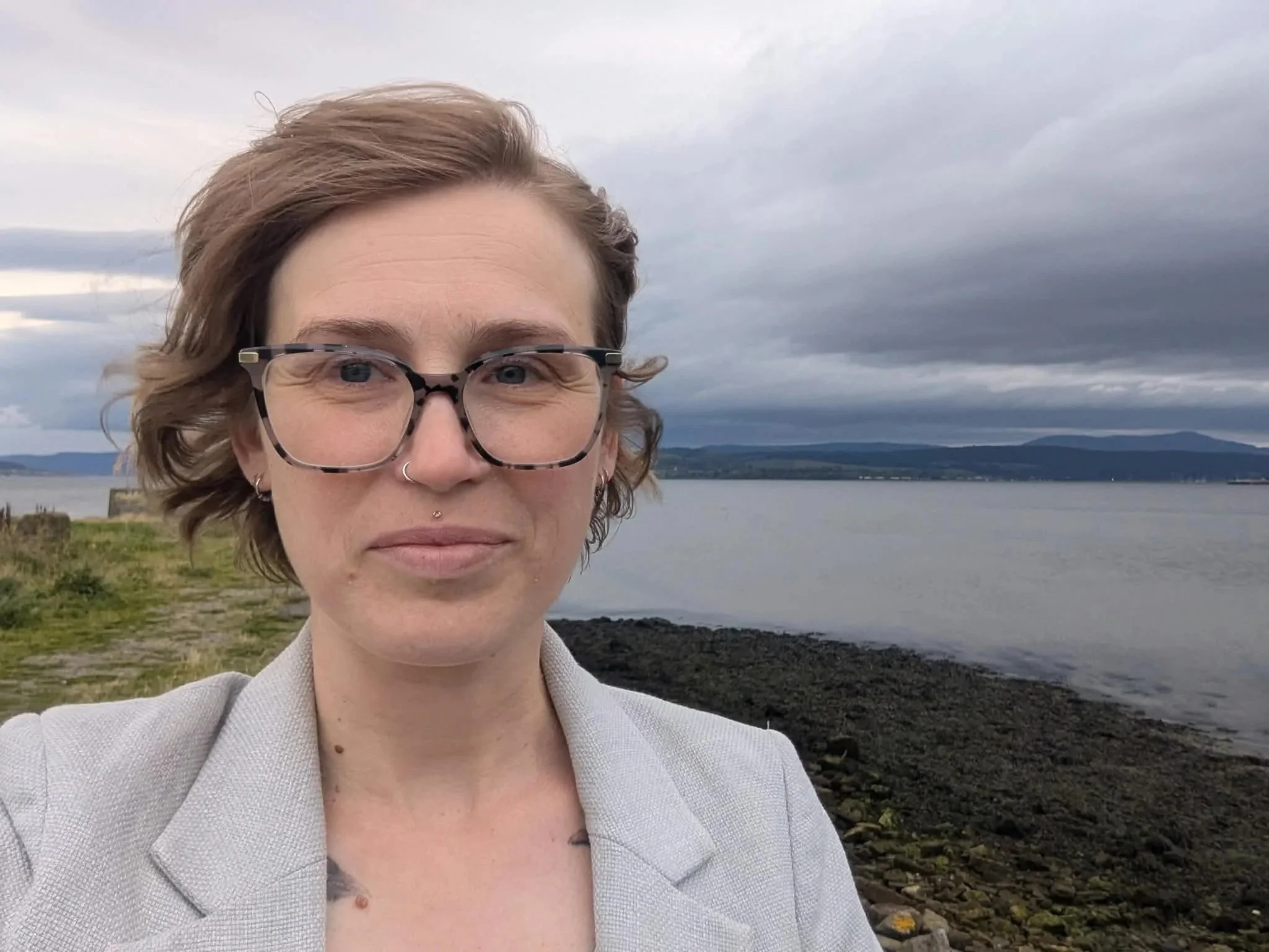 A person with short, wavy hair and glasses, standing near a body of water with a mountainous shoreline in the background, under a cloudy sky.